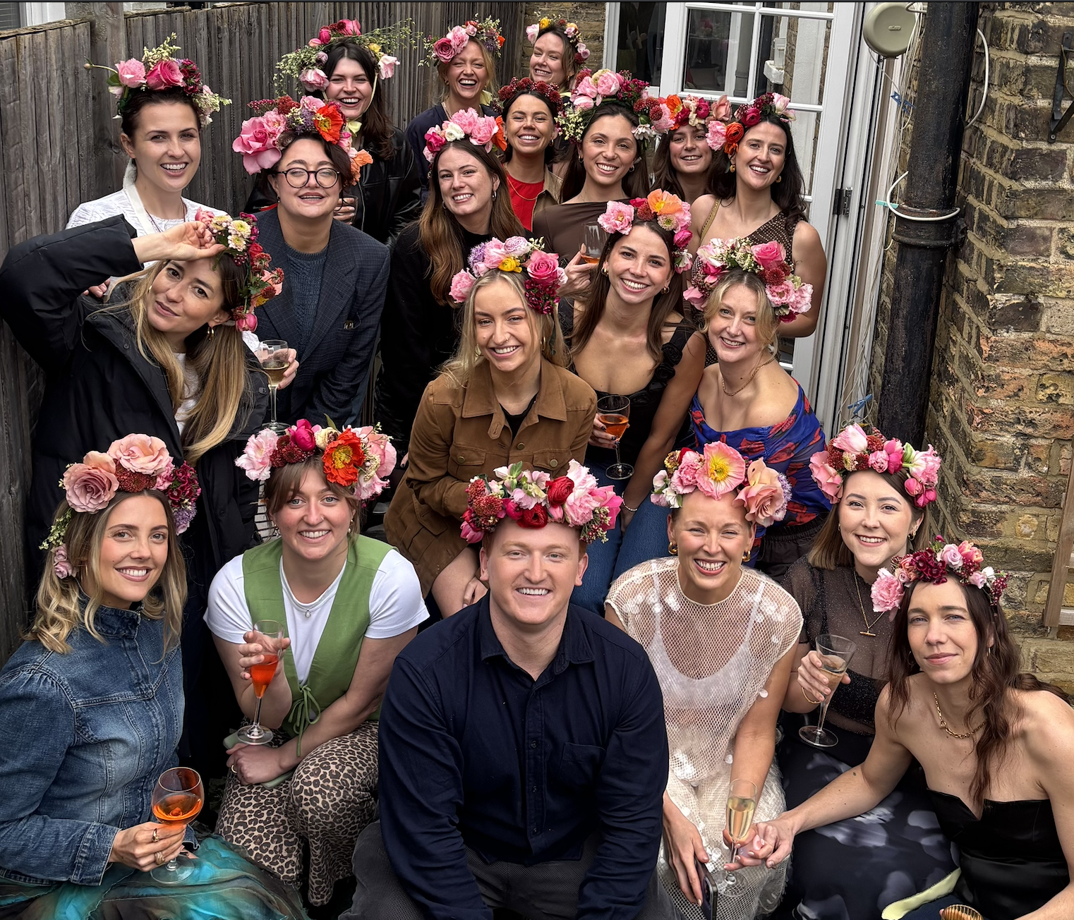 A group of women and one man celebrating outdoors, all wearing flower crowns and holding drinks, smiling and posing together.