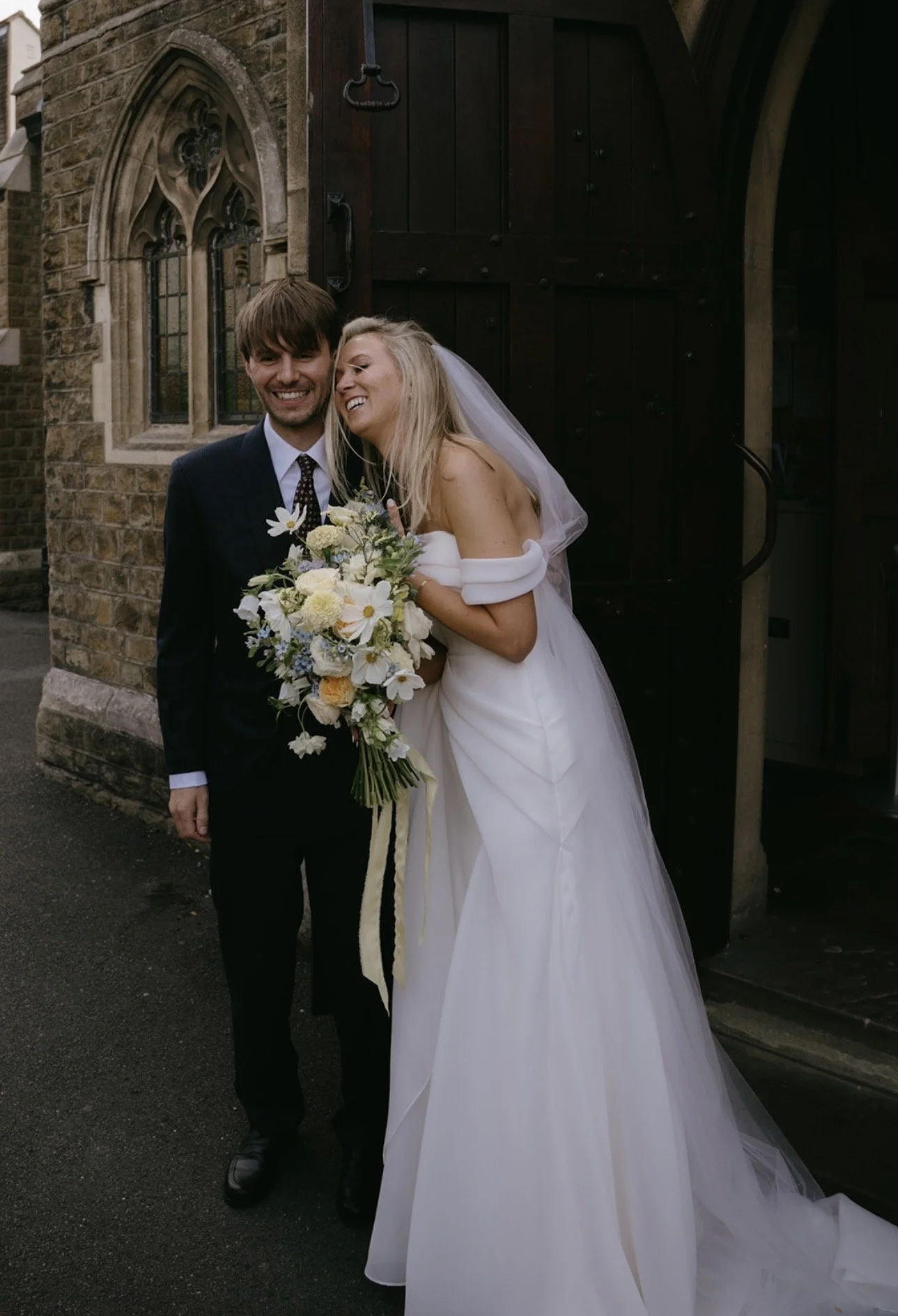 A newlywed couple standing outside a church, smiling and hugging. The bride is holding a bouquet of white and yellow flowers, wearing a white wedding dress and veil. The groom is in a dark suit and tie. They are standing near a large wooden door and stone church wall.