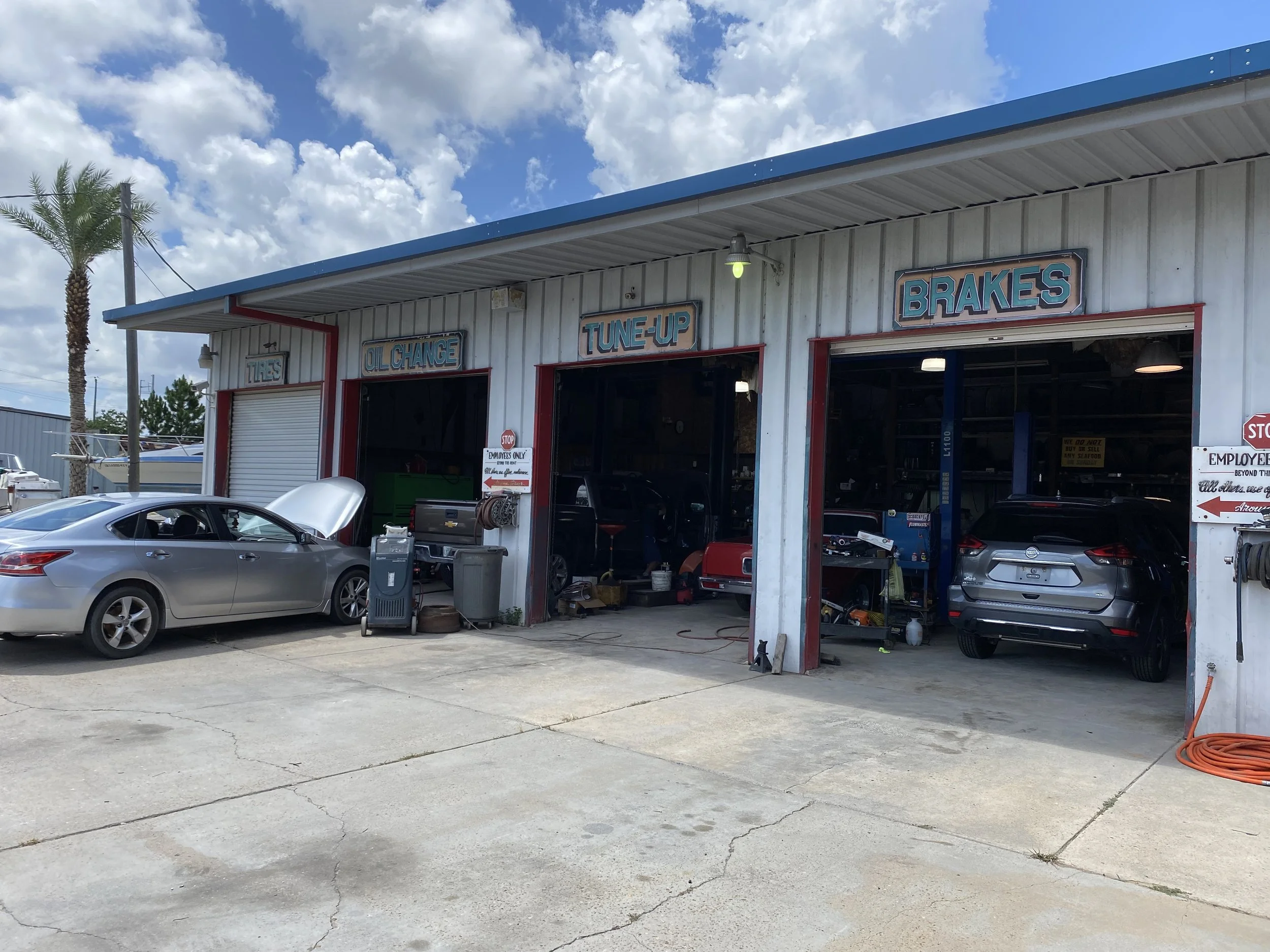 Auto repair shop with signs for tires, oil change, tune-up, and brakes. There are two cars inside and outside the garage, with one car's hood open, and various tools and equipment around.