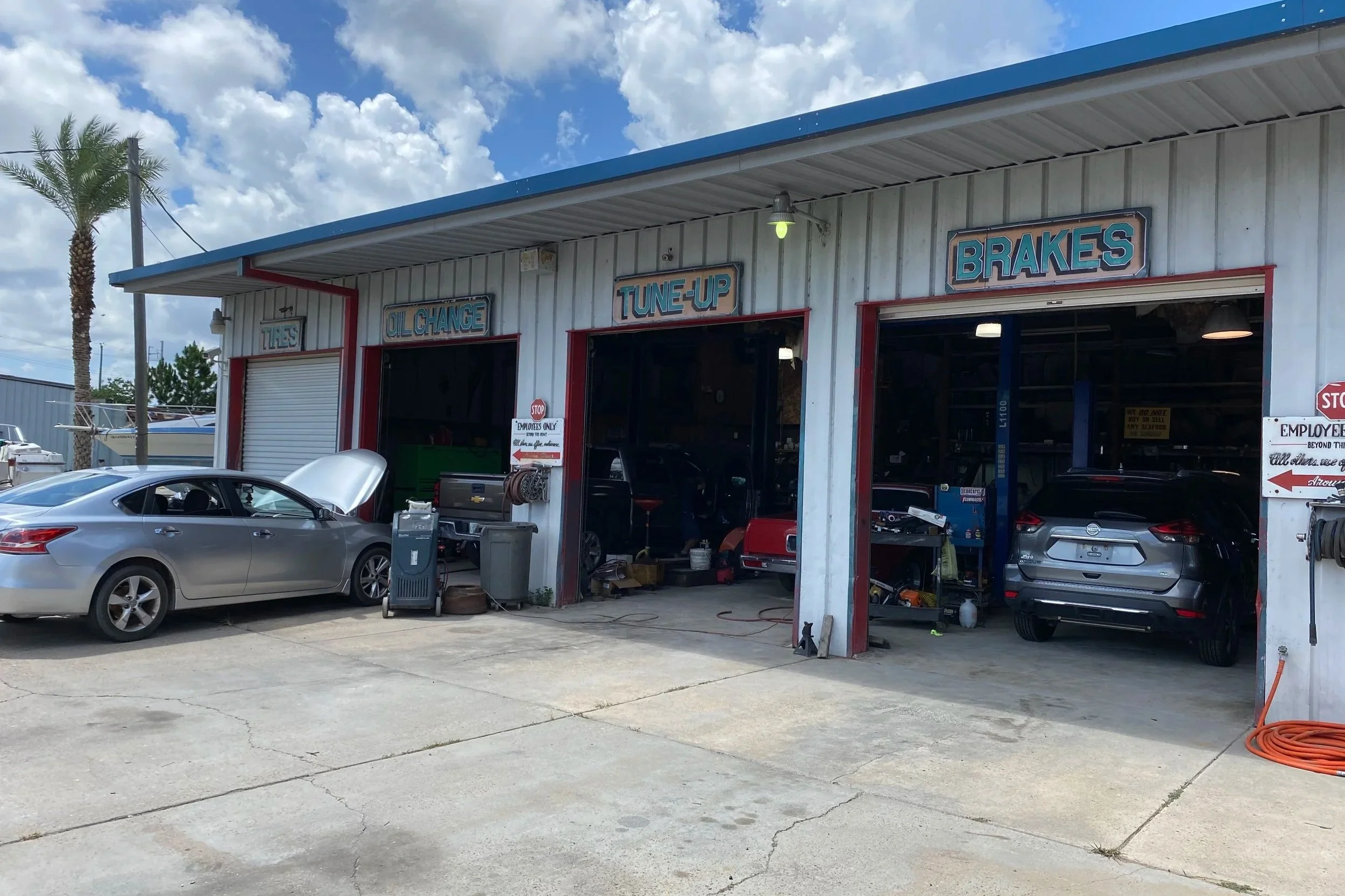 Auto repair shop with three bays, cars being serviced, signs reading 'Tires,' 'Oil Change,' 'Tune-Up,' and 'Brakes,' outdoor parking with a silver sedan and a black SUV, blue sky with clouds.