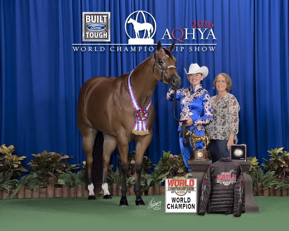 A woman in a blue cowgirl outfit and white hat standing next to a brown horse with a ribbon around its neck, at the 2016 AQHA World Championship Show. The horse is the world champion, and there are various awards and plaques on tables in front of the