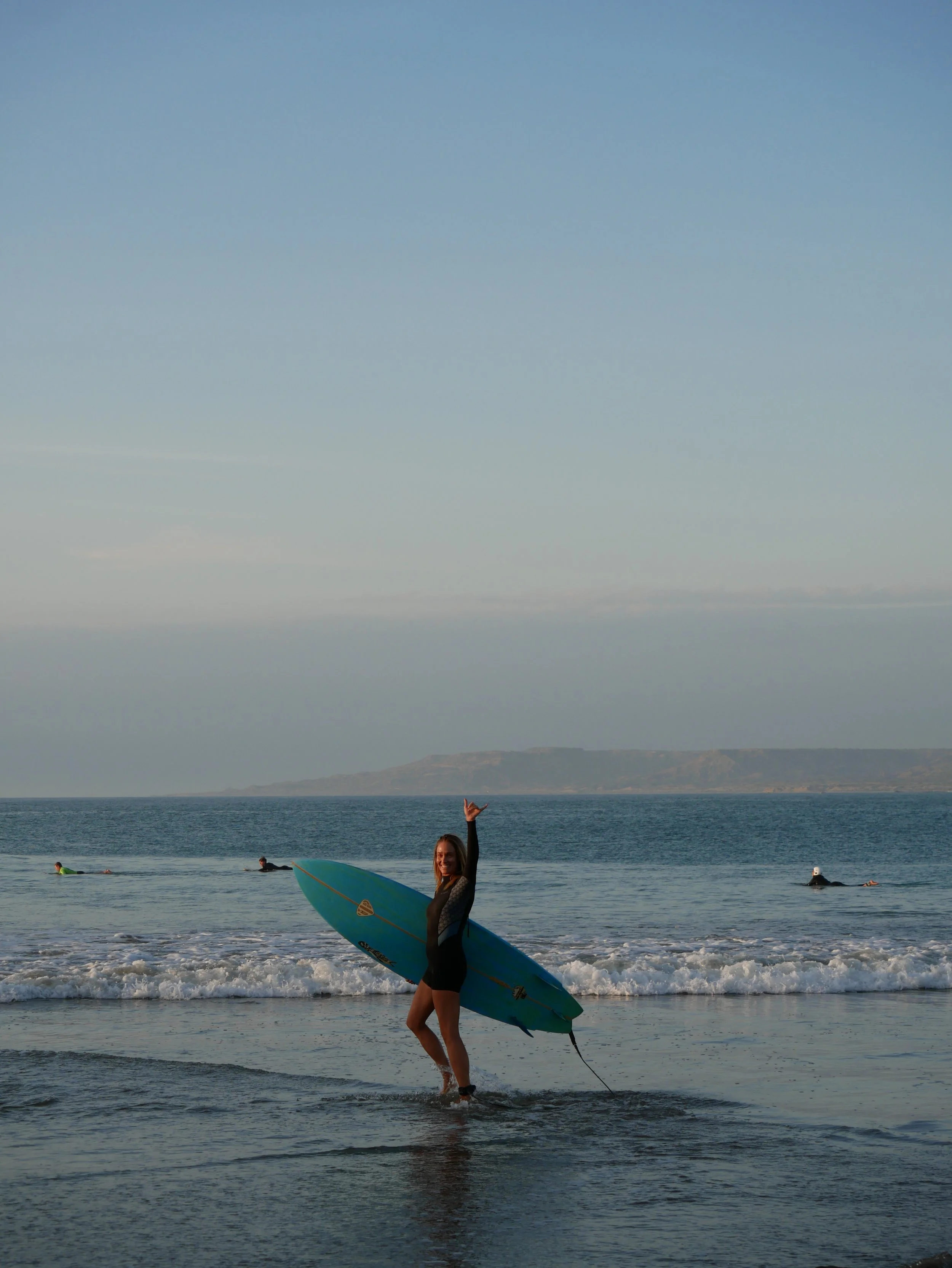 A woman in a wetsuit holding a surfboard in the ocean with coastal houses and cliffs in the background.