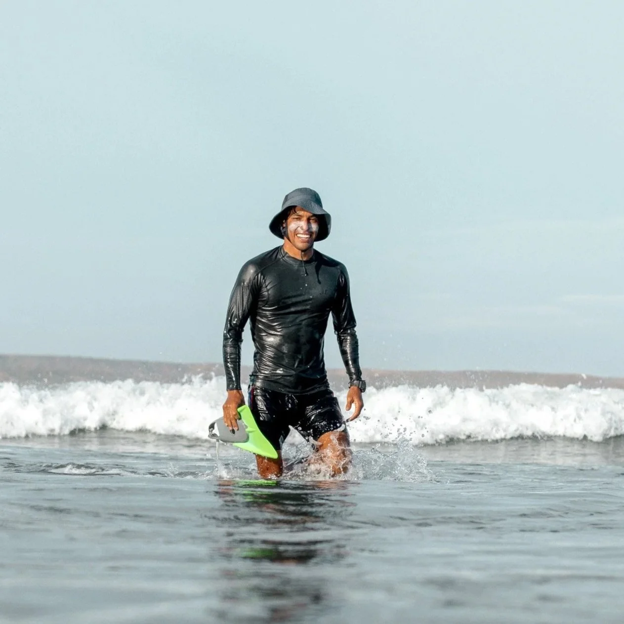 A smiling man in a wetsuit standing in the ocean, holding a cap or hat, with a calm horizon and scattered rocks in the background.