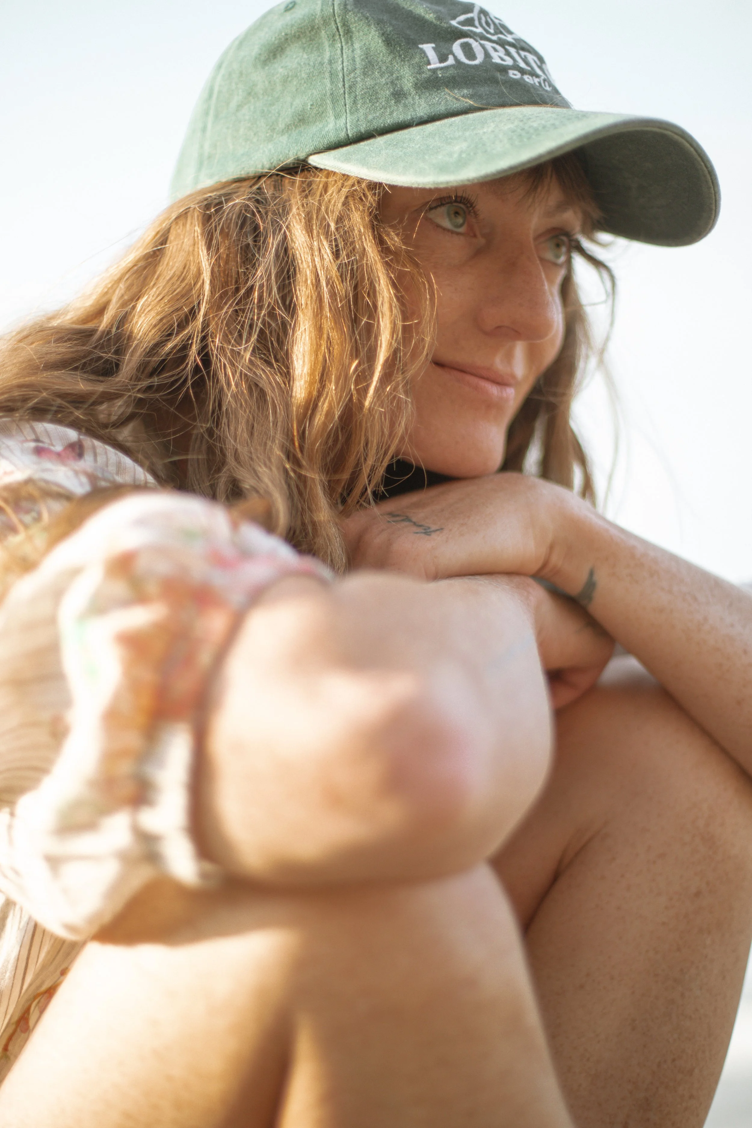 A woman in a wetsuit standing on the beach, holding a surfboard upright next to her. There is sand and a beach hut in the background.