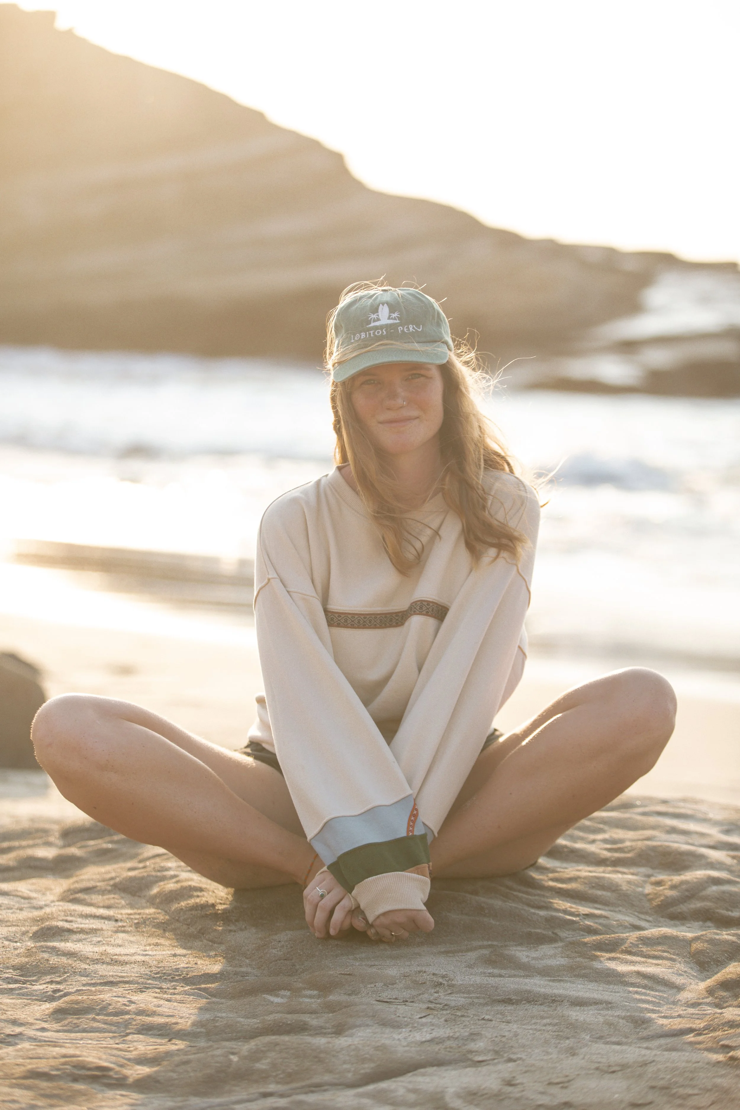 A woman in a hat swimming in the ocean, smiling, with a mountainous landscape in the background.