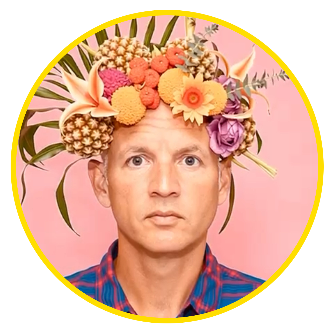 A man with a serious expression wearing a colorful shirt and a flower crown made of various vibrant flowers and greenery, against a pink background.