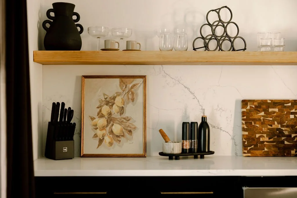 Kitchen shelf with black vase, glassware, artwork of yellow pears, black knives in a holder, spice grinders, and a wooden cutting board.