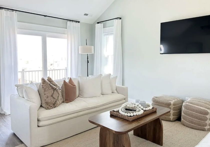 Living room with white sofa, beige and furry pillows, wooden coffee table with decorative items, flat-screen TV mounted on white wall, sunlight coming through from large windows with white curtains, and three beige textured poufs.
