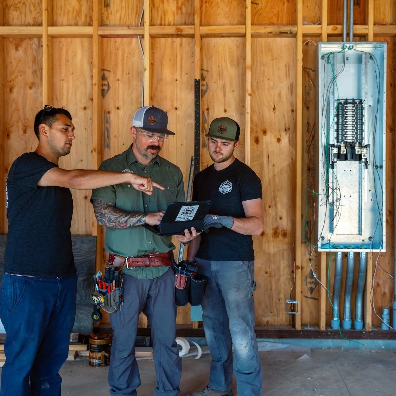 Homeowner and electricians review electrical plans on a laptop beside an open panel during construction, illustrating consultation, panel upgrade assessment, and planning for residential electrical service improvements.