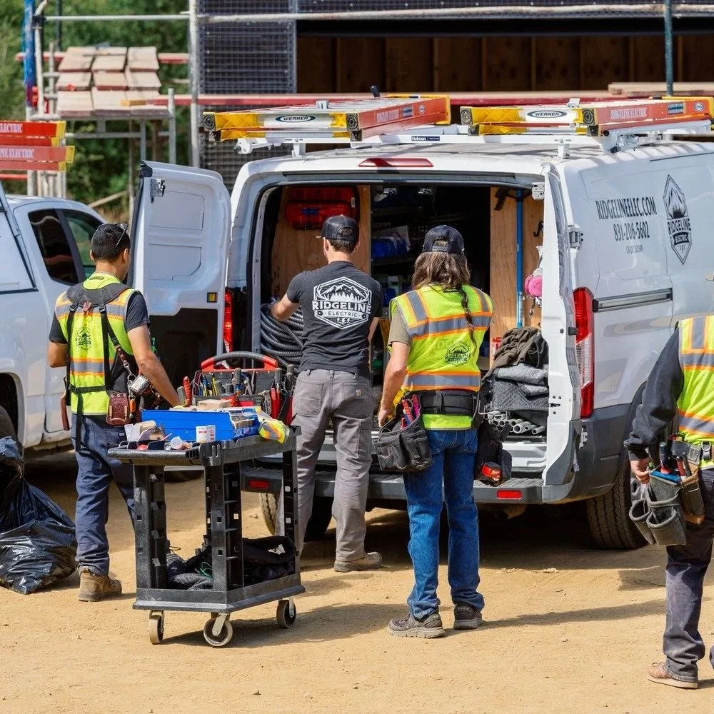 Commercial electricians in safety vests gather around work vans and equipment at a job site, preparing for outdoor lighting installation, maintenance, or electrical infrastructure upgrades on a property.