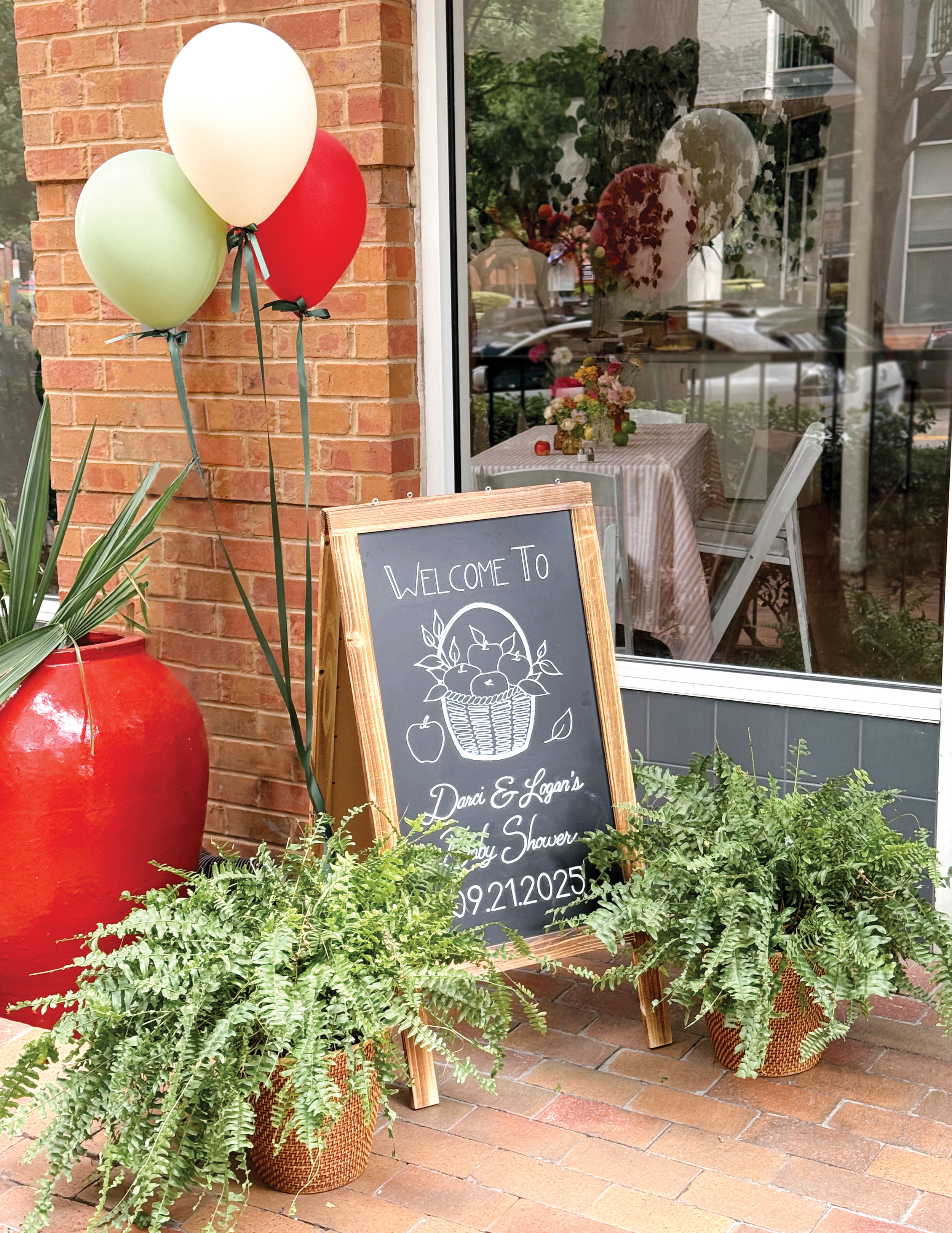 Outdoor storefront with balloons, potted ferns, and a chalkboard sign that reads 'Welcome to Darci & Logan's Baby Shower' with date 9.21.2025. The store has a brick exterior and a window showing a decorated table inside.