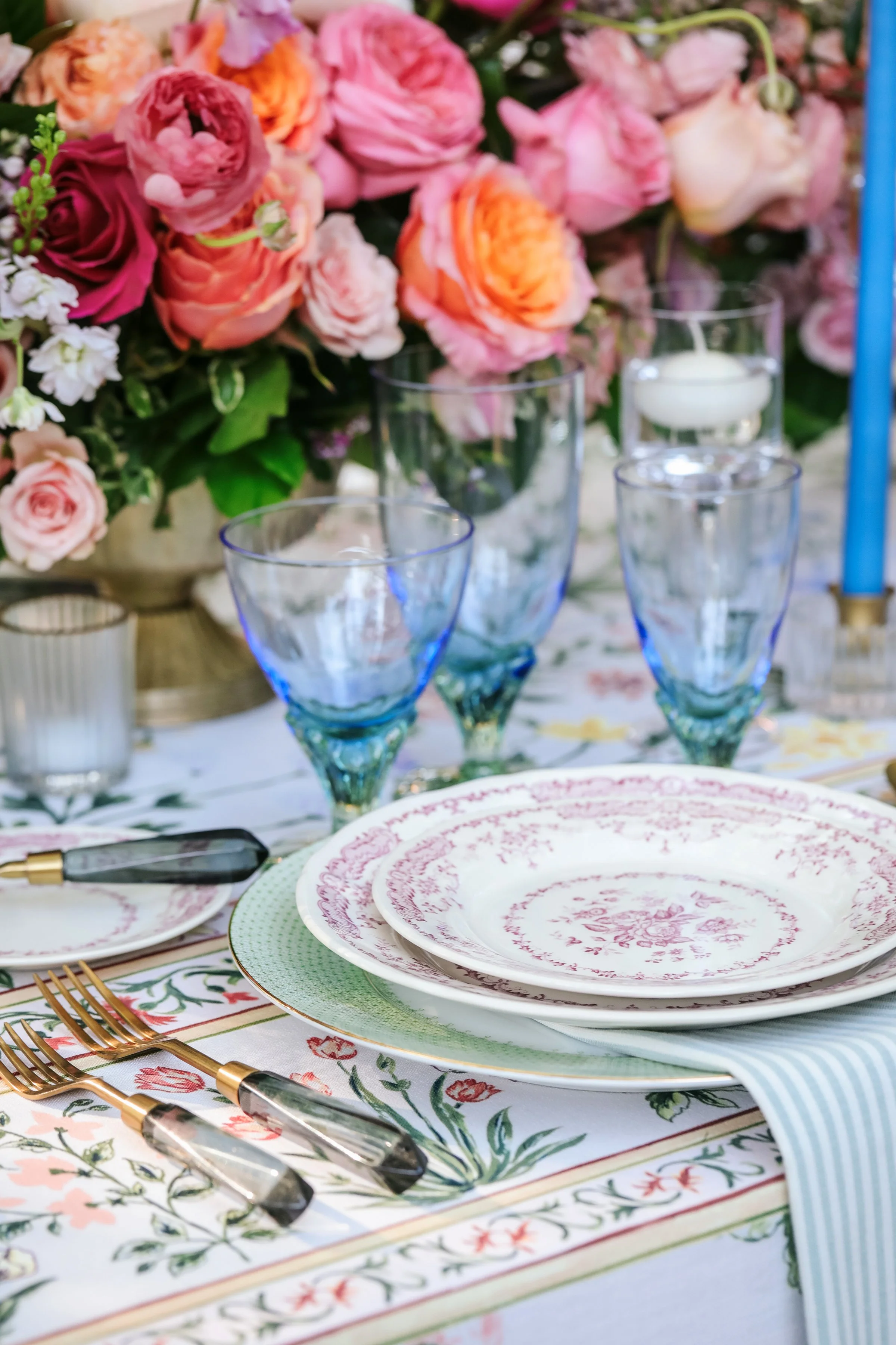 A table setting with pink, orange, and cream flowers in a large vase, arranged among blue-tinted glasses, white plates with pink floral patterns, gold forks with black handles, and a floral tablecloth.