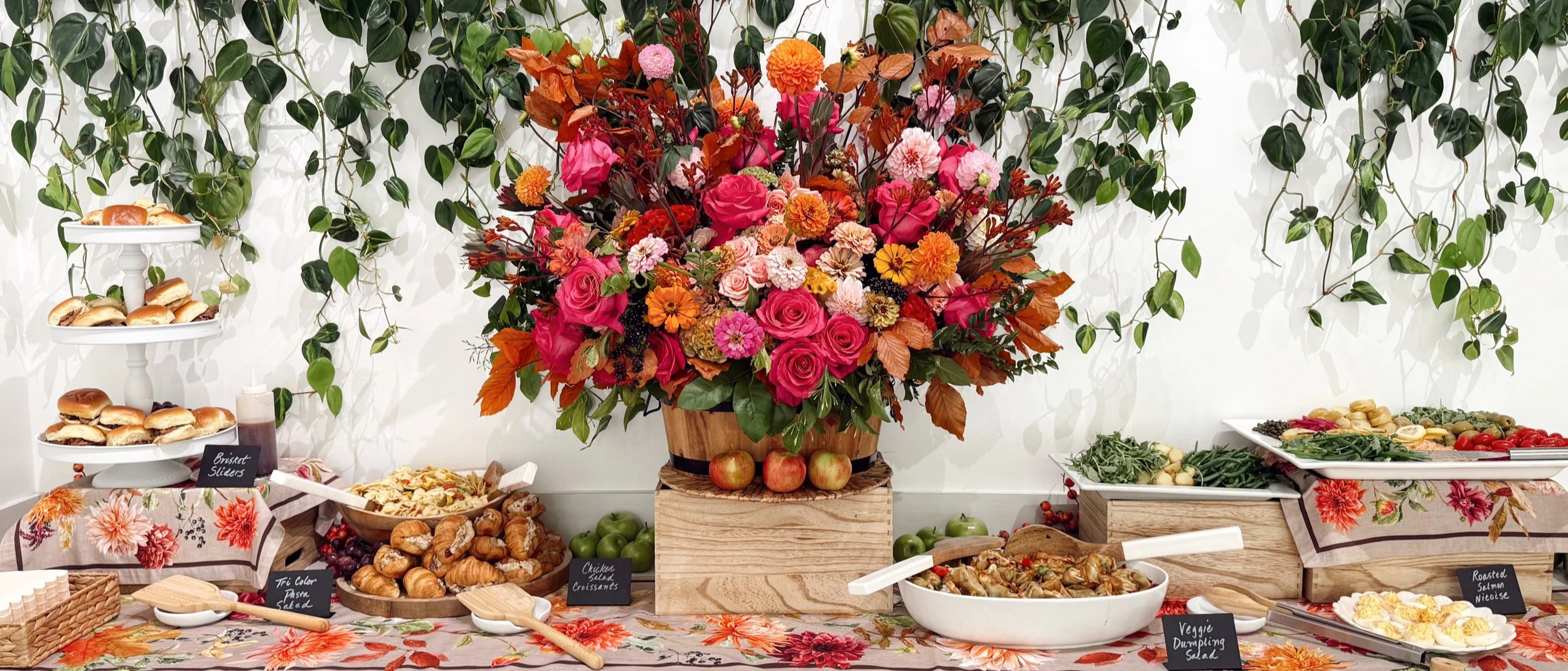 A colorful buffet table with a large floral arrangement in the center, surrounded by various salads, sandwiches, and appetizers, with green leafy vines hanging on the white wall behind.