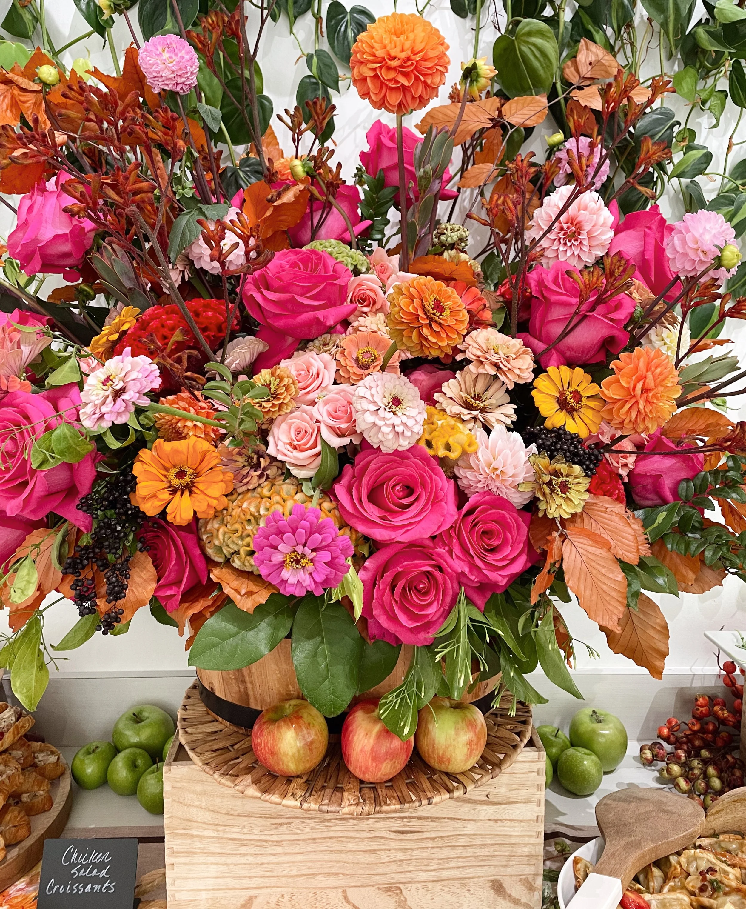 A vibrant bouquet of pink, orange, and peach flowers, including roses and dahlias, arranged in a wooden container on a wicker tray. Apples and greenery surround the arrangement.