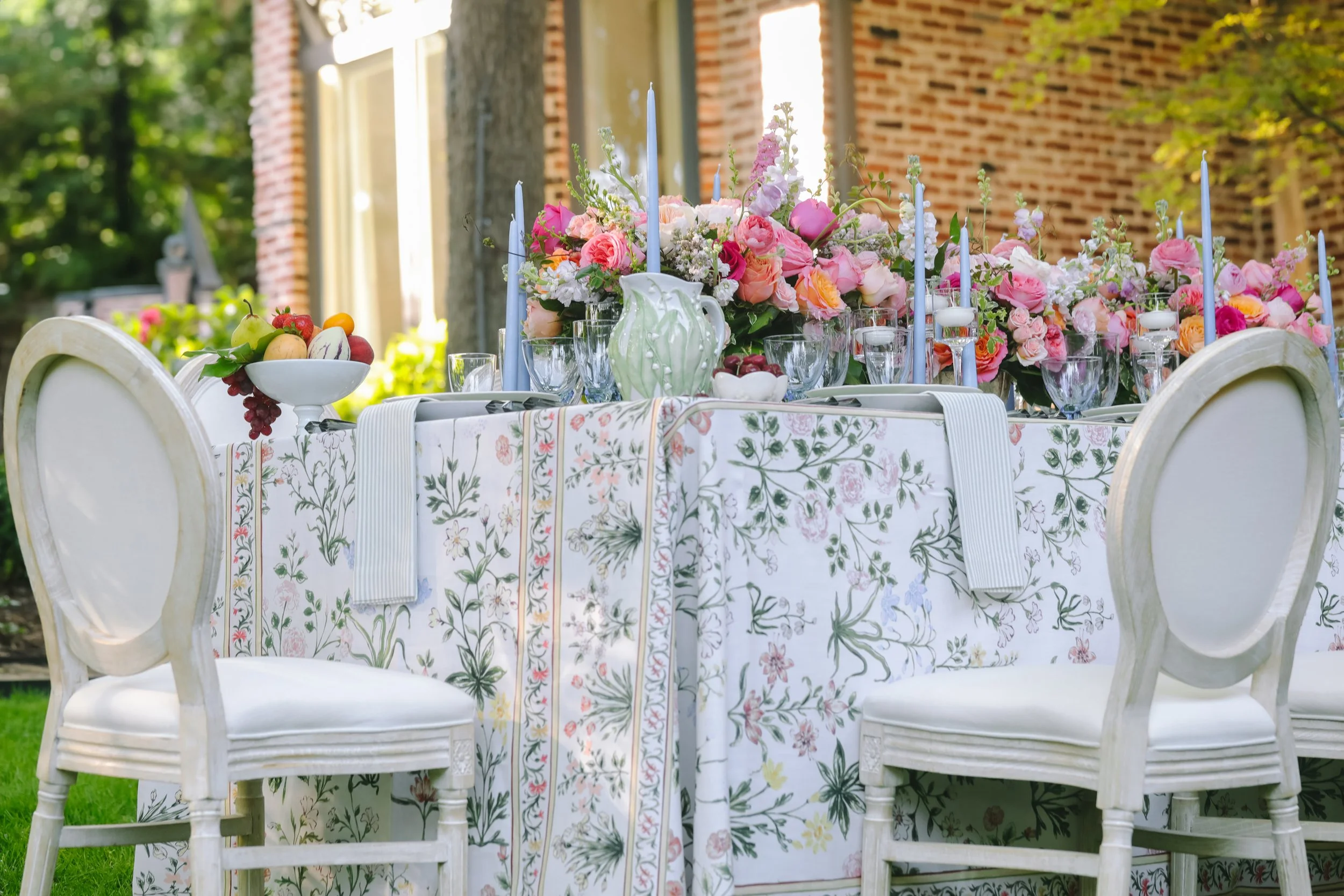 A beautifully decorated outdoor table with floral tablecloth, pink and white floral centerpiece, glassware, blue candles, and a bowl of fruit. White chairs with rounded backs surround the table.