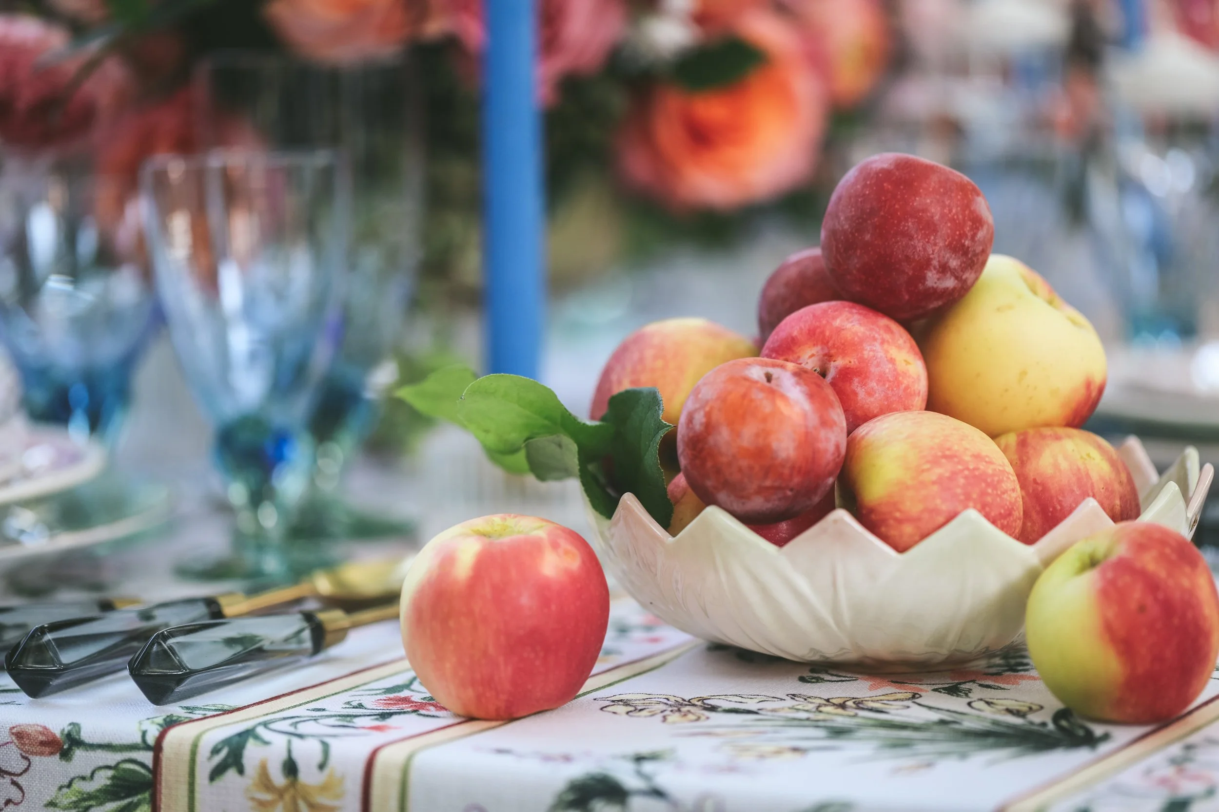 A bowl of apples and grapes on a table with floral tablecloth, with cutlery and glasses in the background.