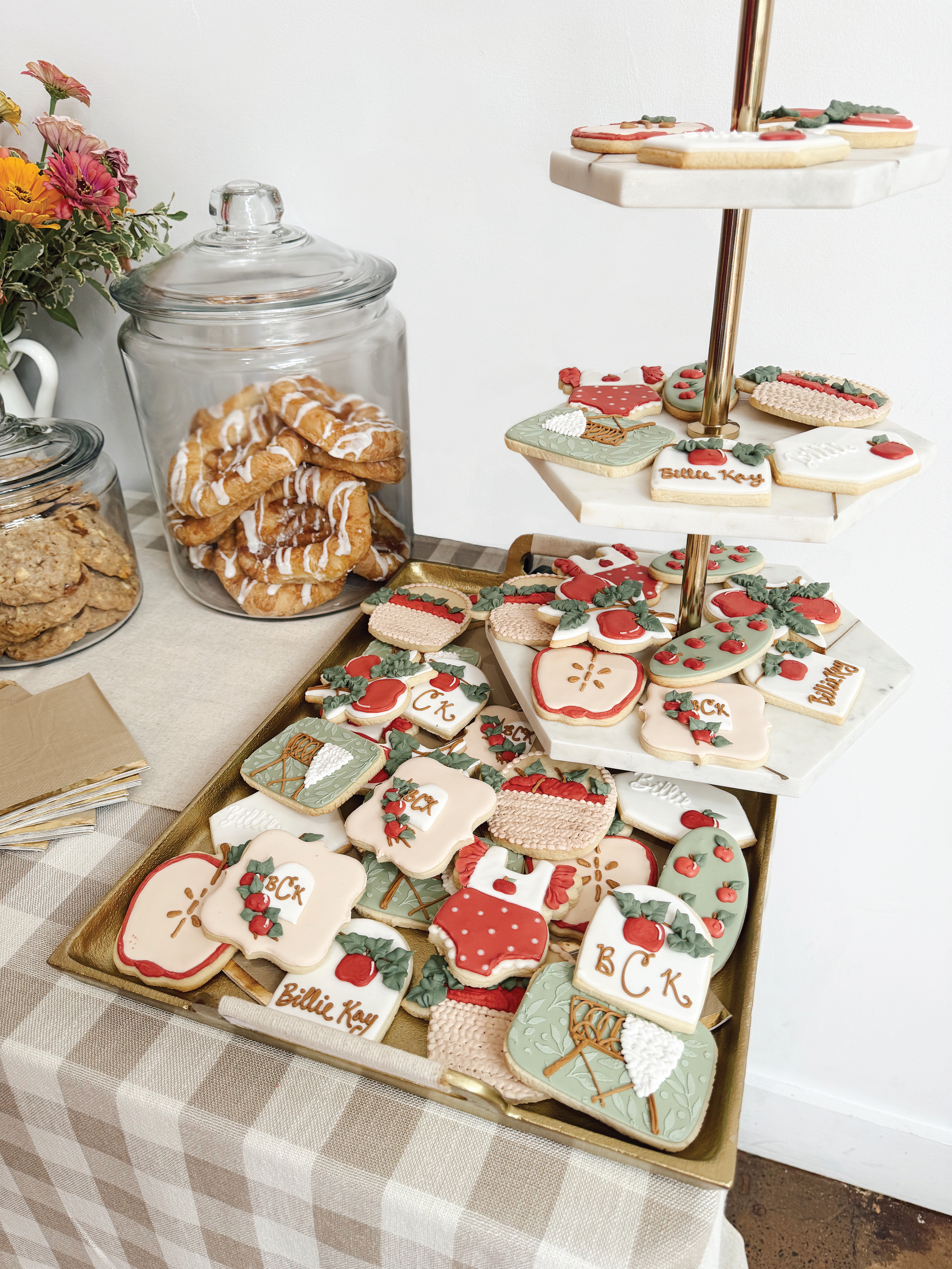 A table with decorated Christmas cookies and jars of cookies, featuring a tiered stand of holiday-themed cookies, a jar of biscotti, and a jar of frosted pastries with a nearby vase of flowers.