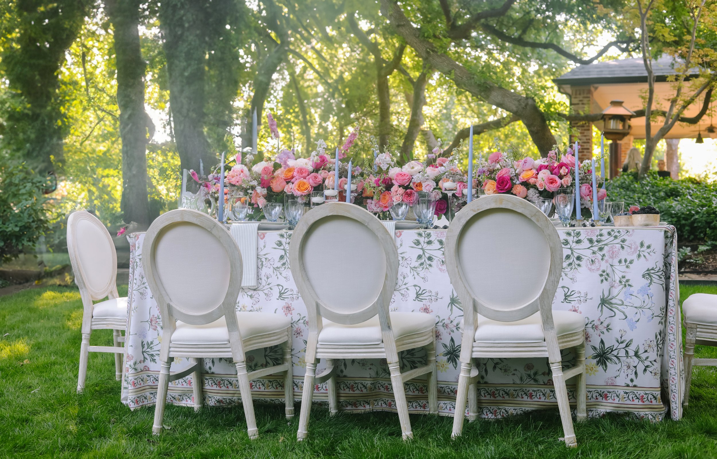 Elegant outdoor table setup with floral centerpiece, surrounded by white chairs, set in a garden with trees and a gazebo in the background.