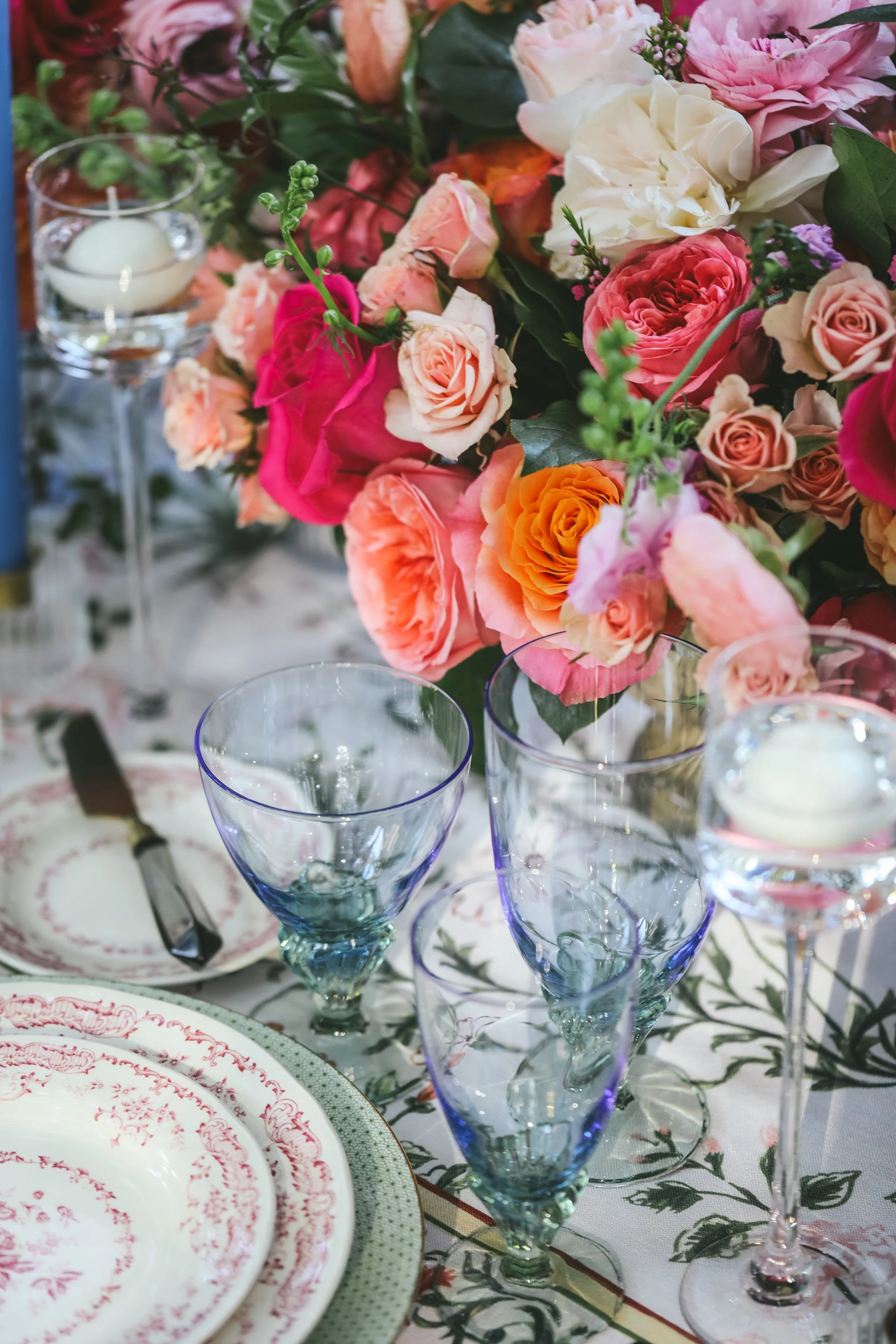 Elegant table setting with floral centerpiece featuring pink, orange, and white roses, surrounded by crystal glasses, decorative plates with red floral patterns, and a tablecloth with a green and white floral design.