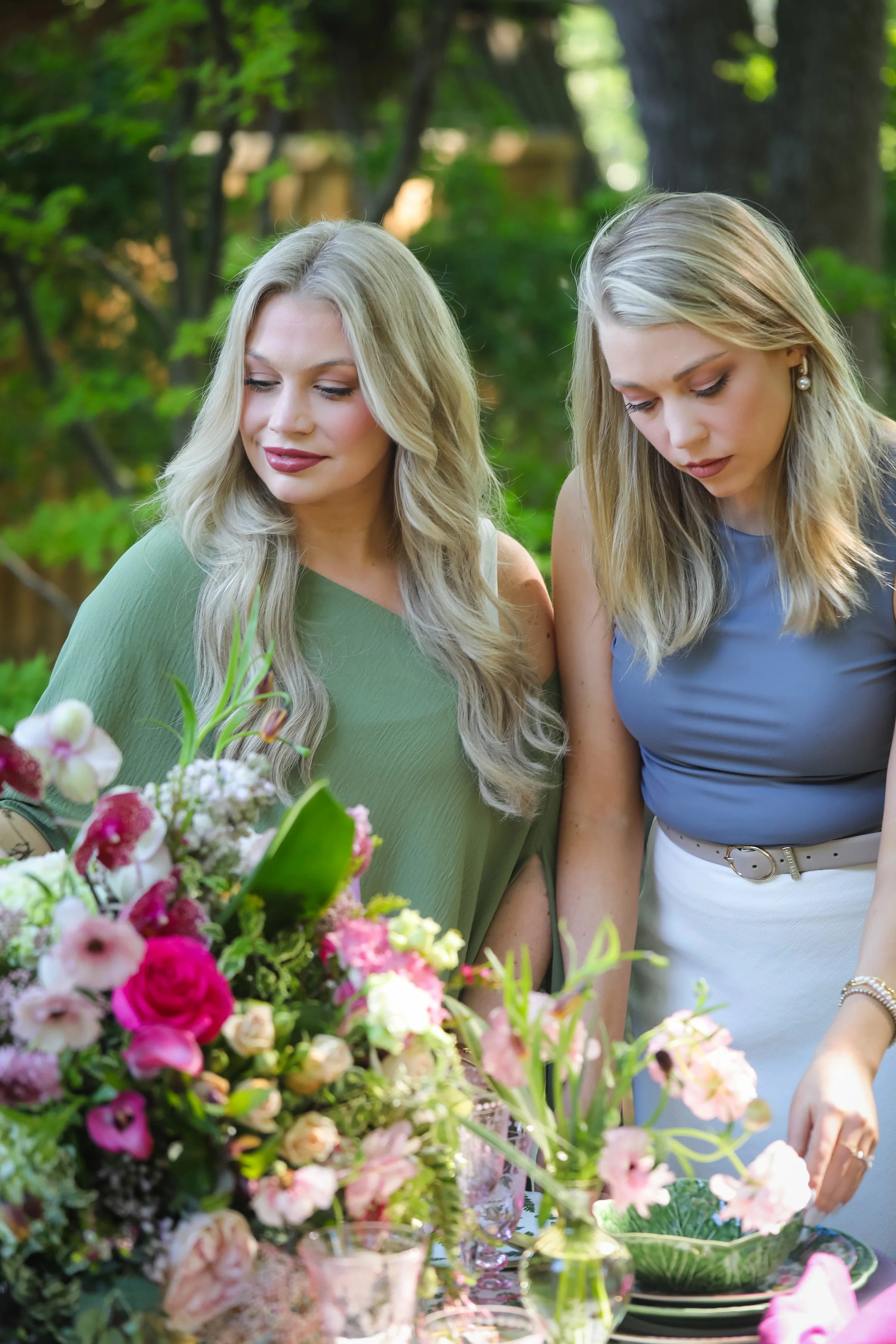 Two women standing outdoors, arranging a colorful floral centerpiece on a table, surrounded by greenery.