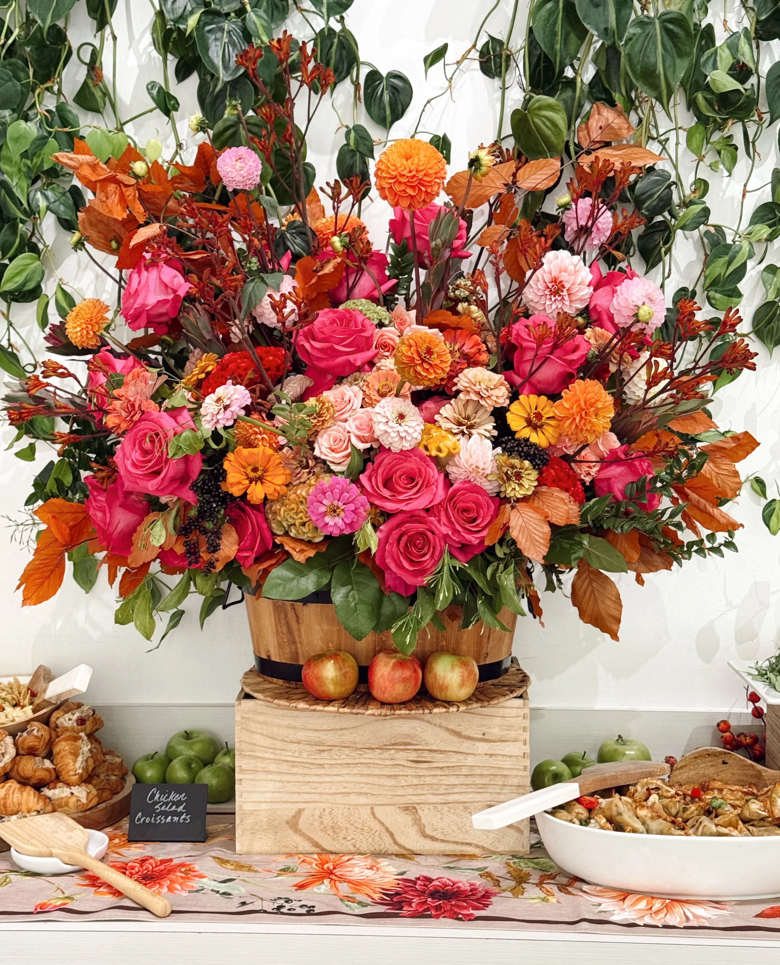 A large arrangement of pink, orange, and peach flowers, including roses and dahlias, in a wooden container on a table with apples and food.