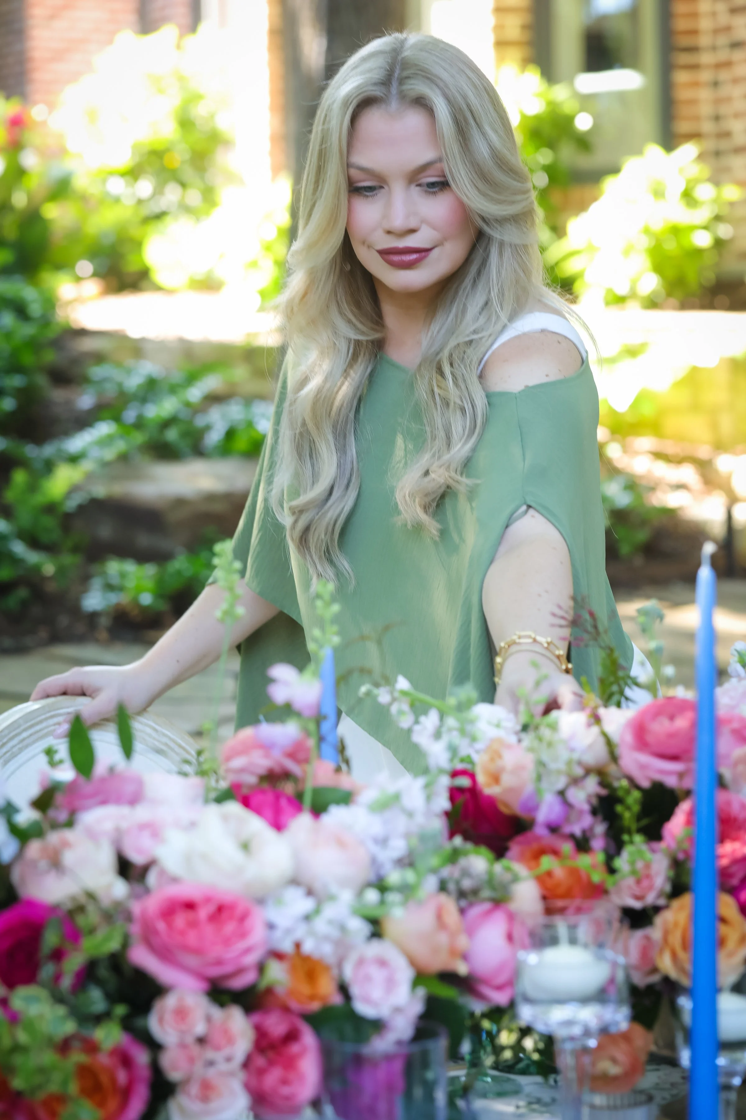A woman with long blonde hair and a green top arranging flowers and candles on an outdoor table decorated with colorful flowers.