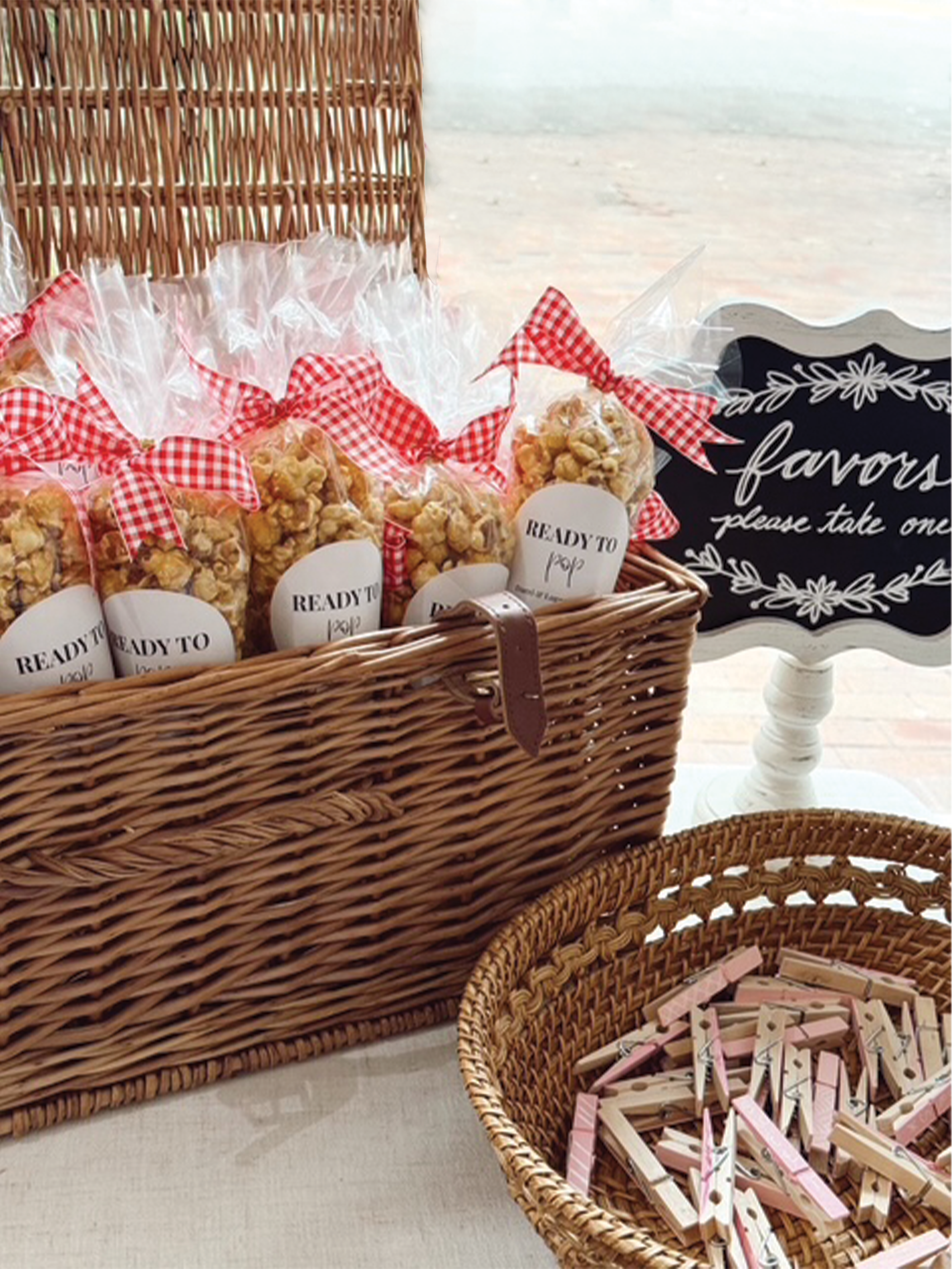A wicker picnic basket filled with individually wrapped popcorn bags with red and white checkered ribbons, a chalkboard sign that says 'favors, please take one,' and a wicker basket containing pink and white clothespins.