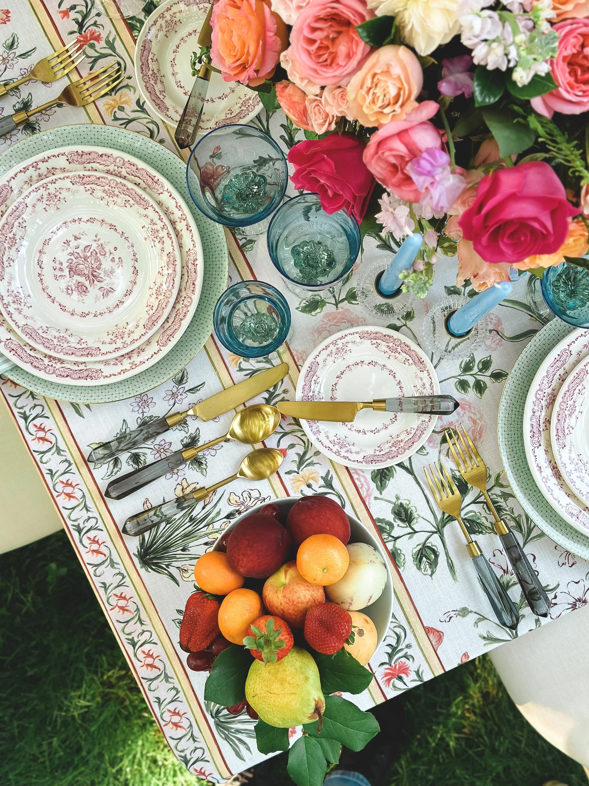 A beautifully set outdoor dining table with floral plates, gold flatware, blue glasses, and a centerpiece of pink, peach, and white roses. There is a fruit bowl with apples, strawberries, peaches, and other fruits, and the table has a floral patterne