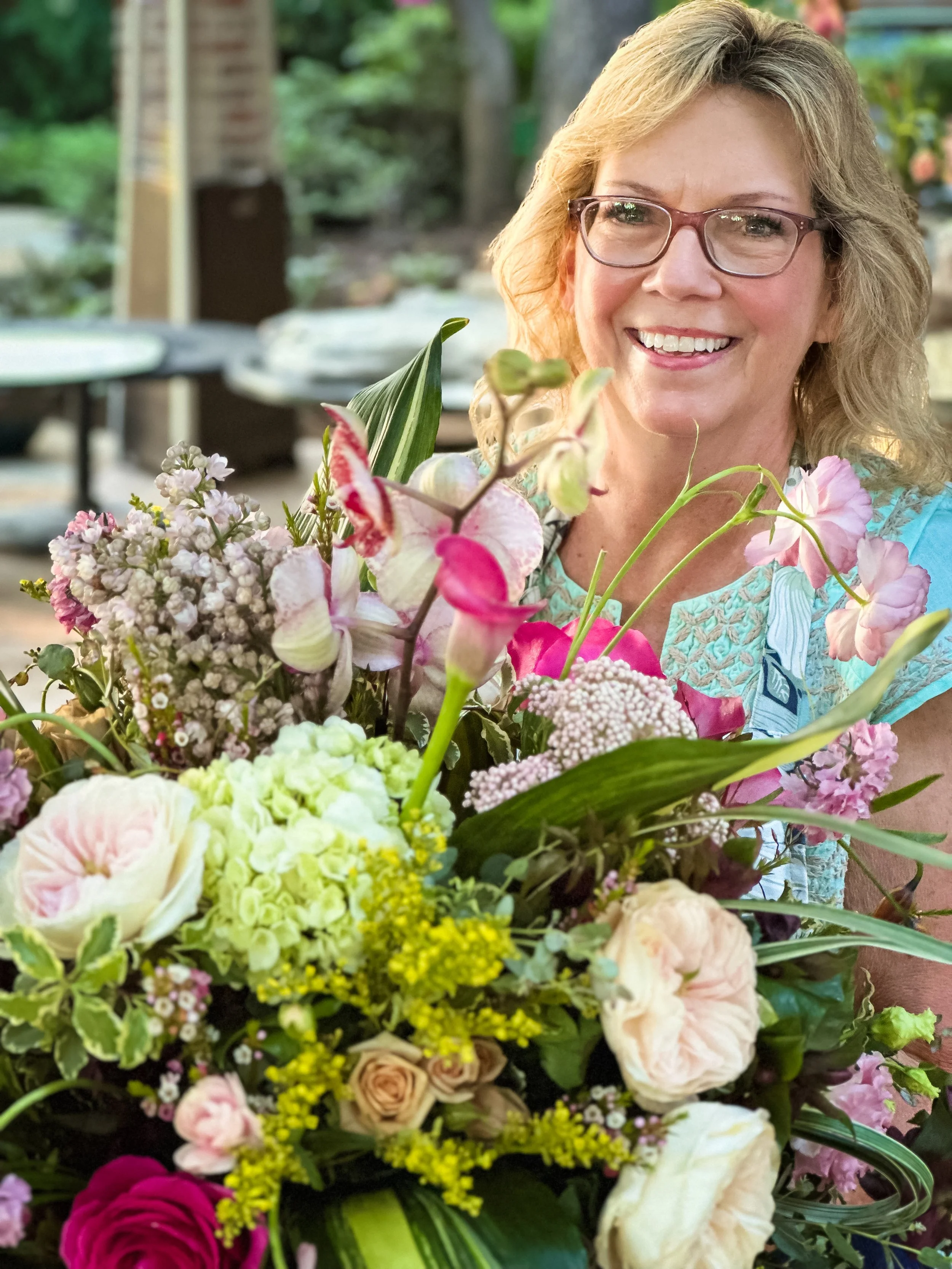 A smiling woman with glasses and blonde hair holding a large bouquet of various colorful flowers.