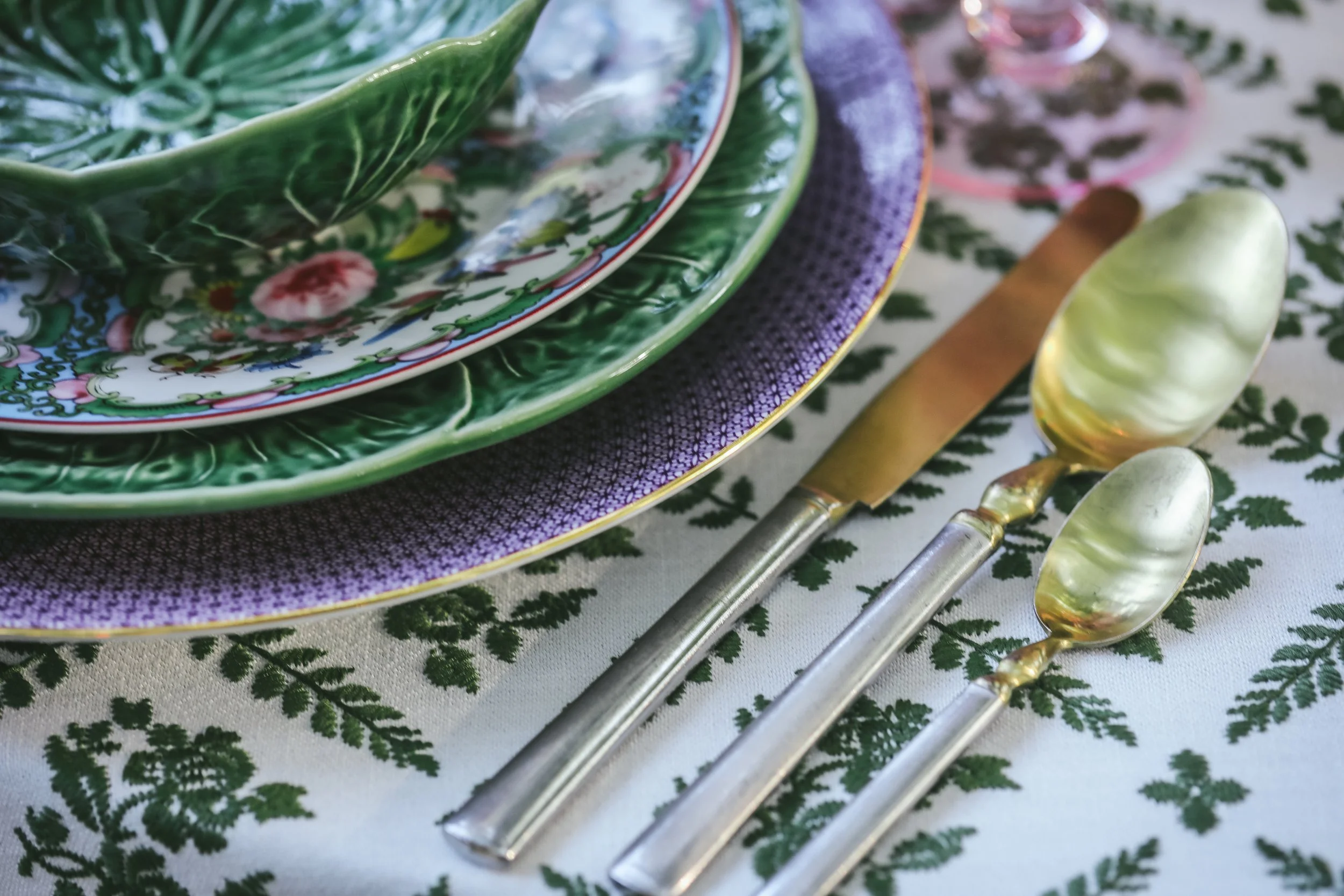 A set of plates, a knife, and two spoons arranged on a decorative tablecloth. The plates are nested, with the top plate featuring a green leafy design, a floral pattern, and a red and white border. The tablecloth has a green leafy pattern, and the ut