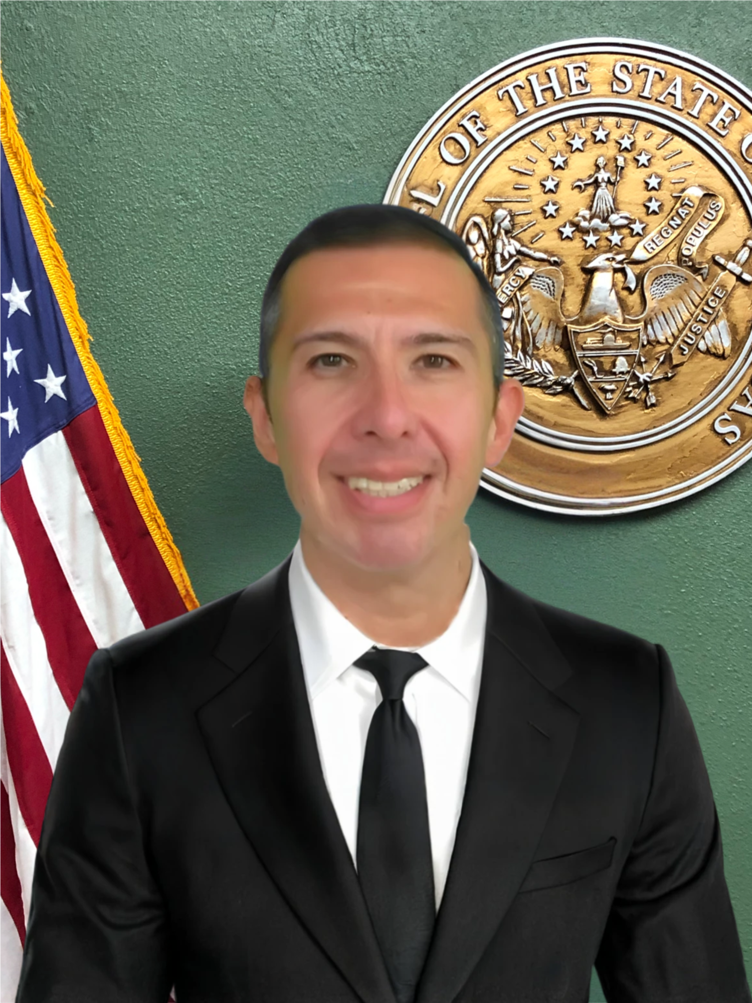 A man in a black suit, white shirt, and black tie standing in front of a green wall with a large bronze-colored state seal and an American flag visible in the background.
