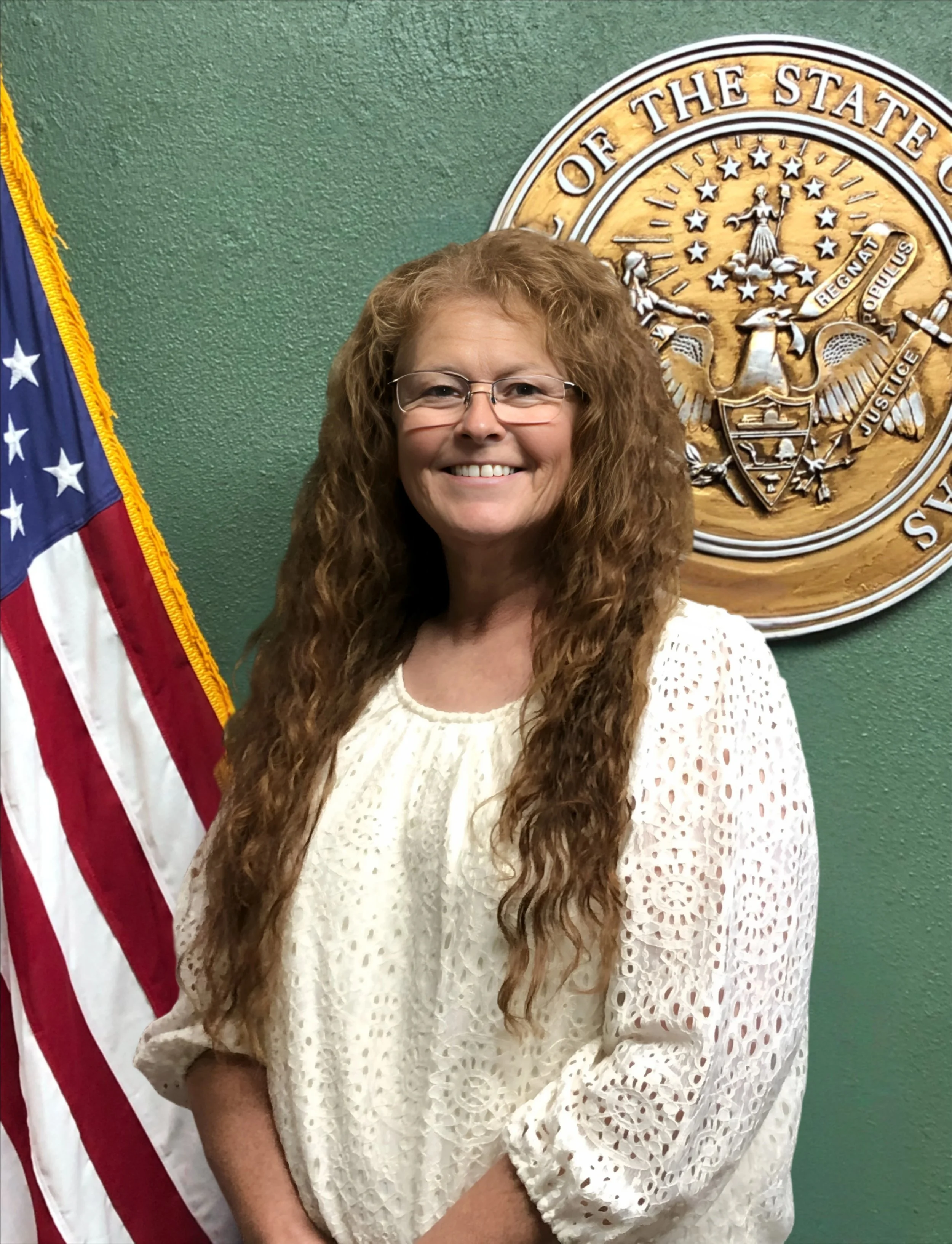 A woman with long, curly red hair and glasses smiling, standing in front of an American flag and a wooden state seal on a green wall.