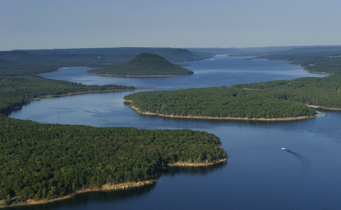 Aerial view of Greers Ferry Lake amid lush green landscape and distant hills under clear blue sky.