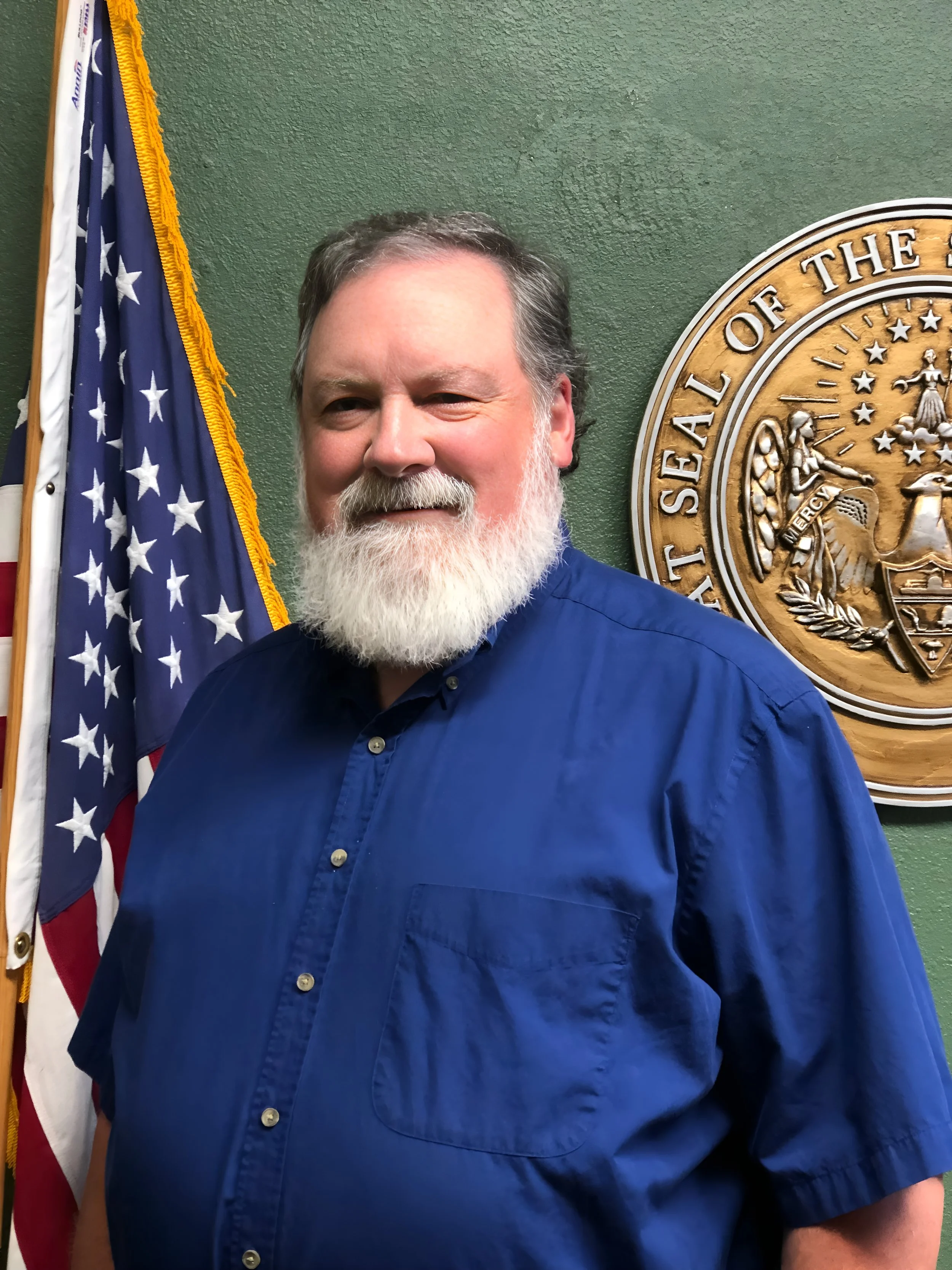 A smiling man with a white beard in a blue shirt standing in front of an American flag and a seal that reads "State Seal of the".