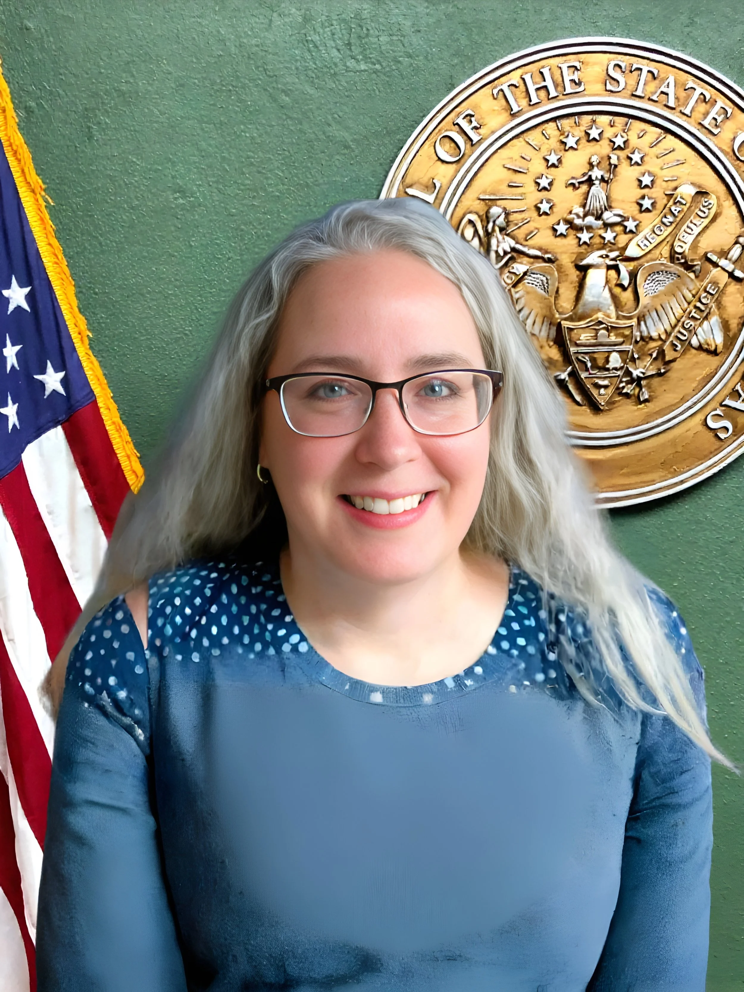 A woman with glasses and long blonde hair wearing a navy blue dress with white dotted shoulder details, smiling, standing in front of an American flag and a large gold seal of the state of Oregon.