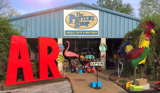 The Pottery Shop storefront with large red 'ART' sculptures outside. Decor includes a pink flamingo, a large colorful rooster sculpture, and various outdoor decorations and gift items visible through the windows.