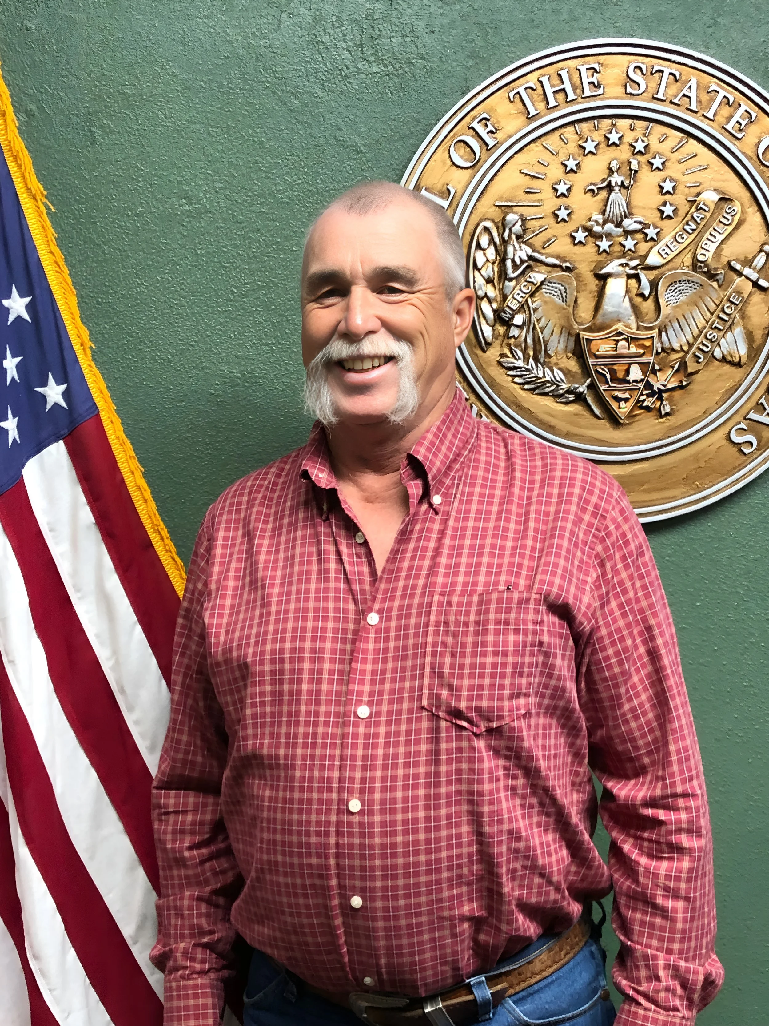 A man with a beard and mustache standing in front of an American flag and a state seal, smiling at the camera.