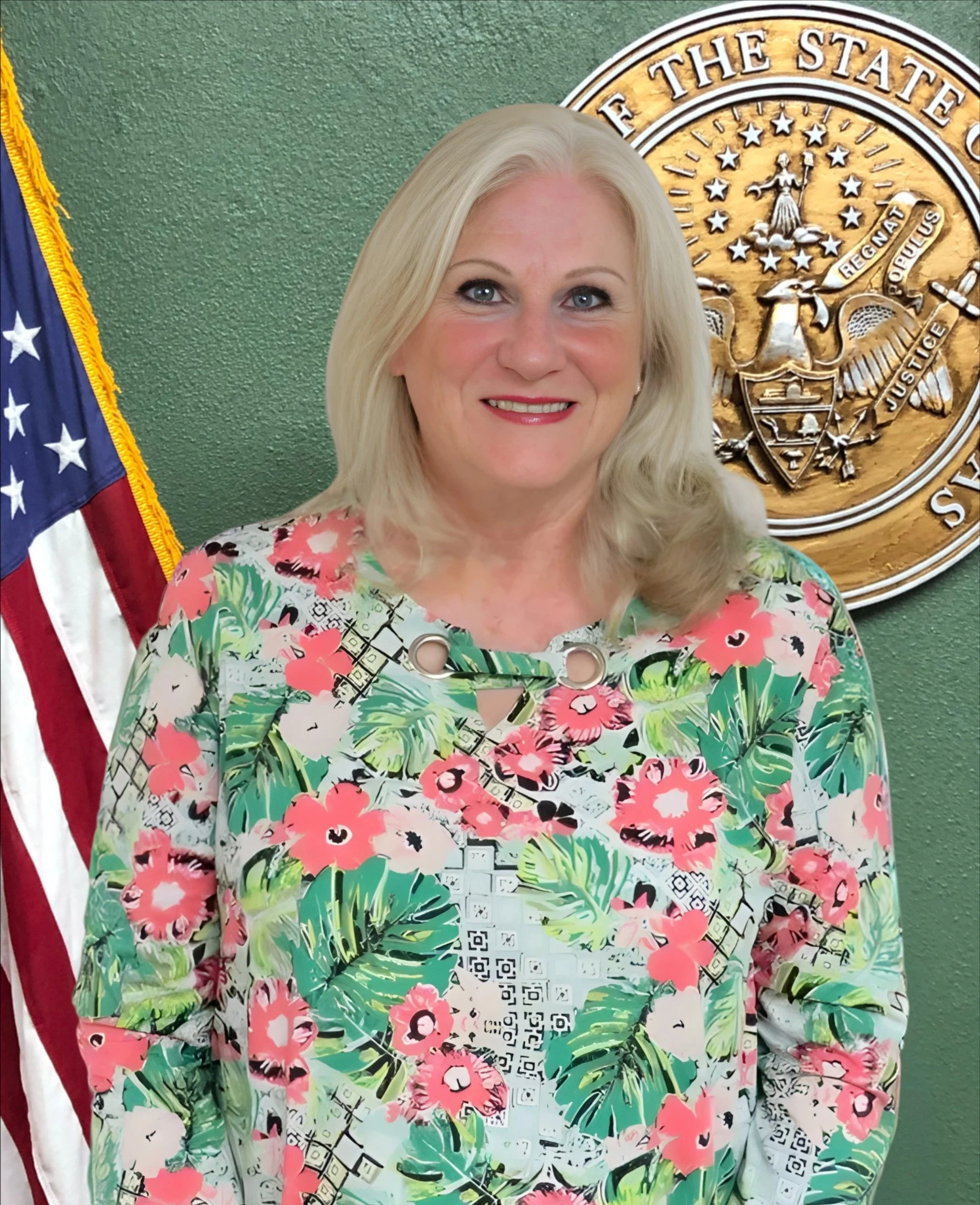 A woman with blonde hair wearing a colorful floral-patterned top, standing in front of a green wall with a state seal and an American flag.