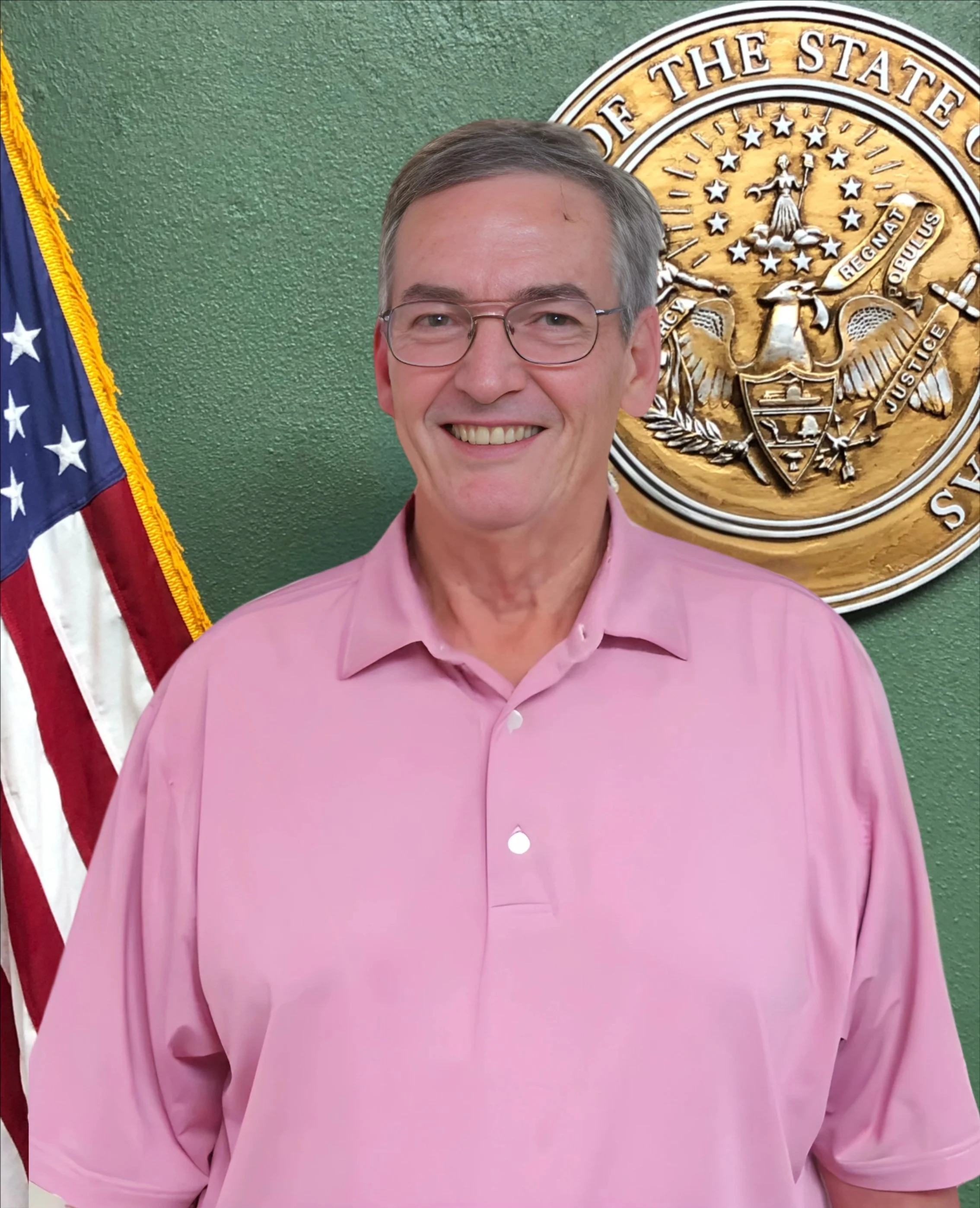 A man in a pink shirt smiling, standing in front of a green wall with a large gold official seal of the state of Georgia and a partial American flag to his left.