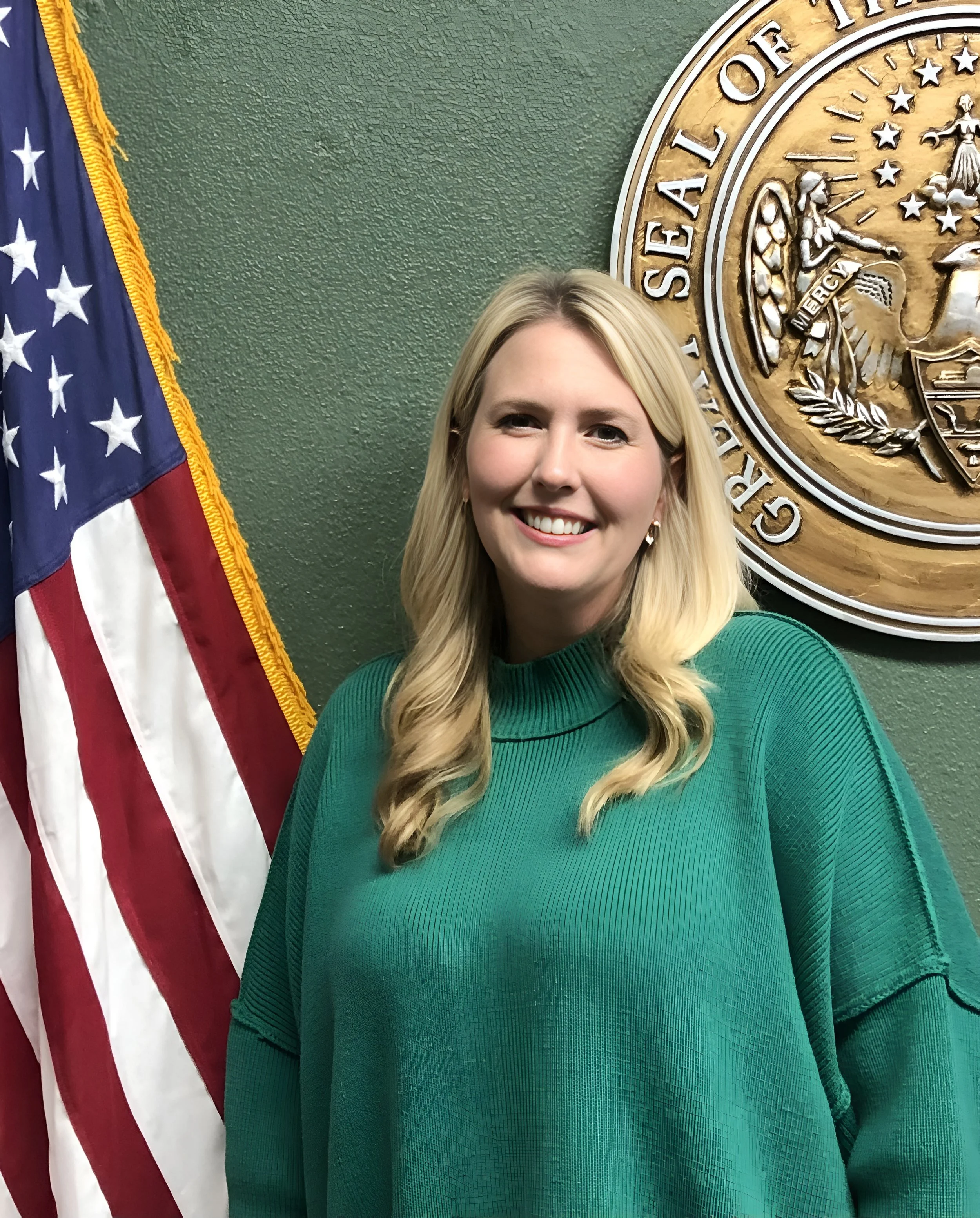 A woman with blonde hair, wearing a green sweater, standing in front of a green wall with a large seal and American flag behind her.