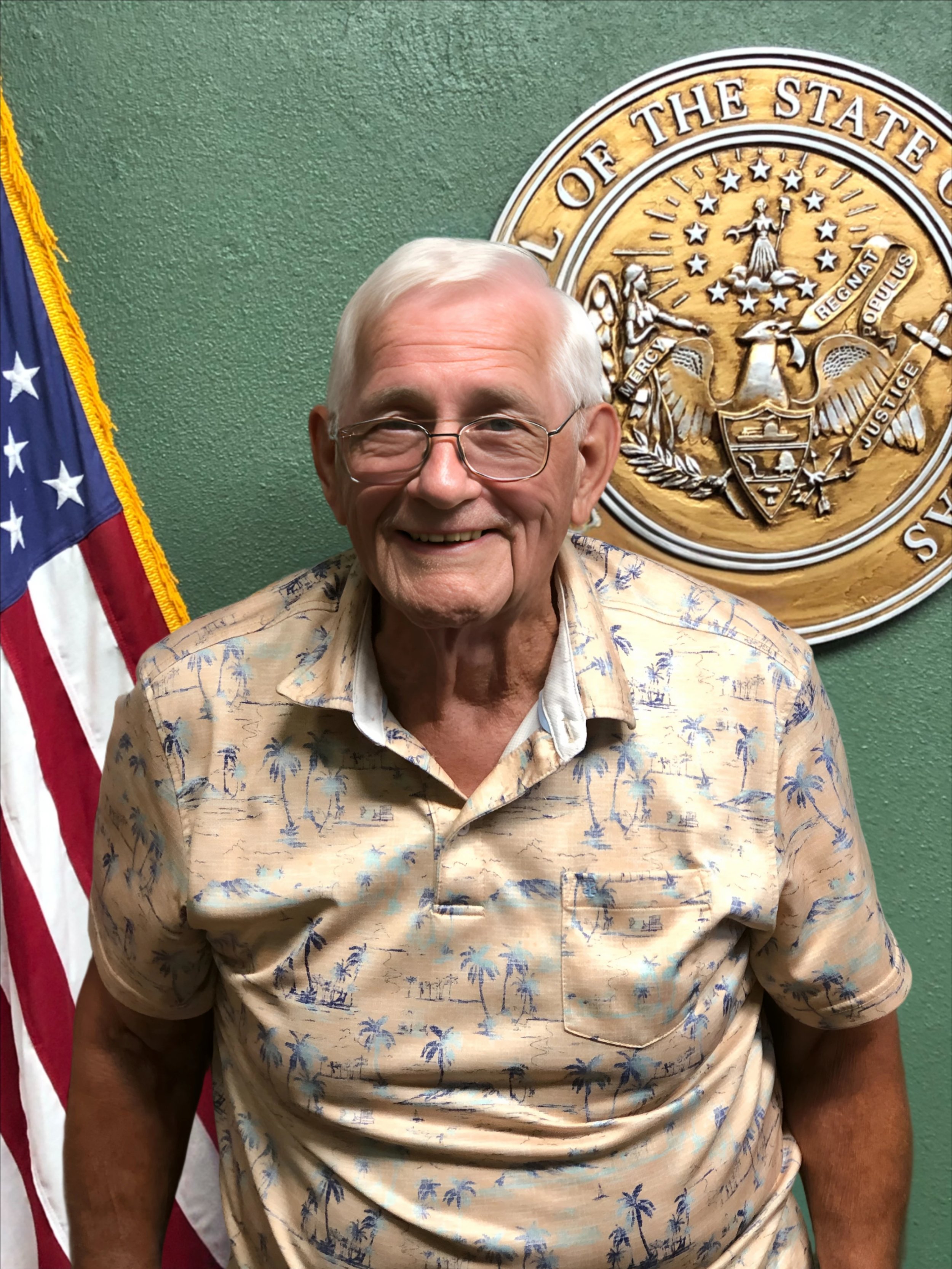 An  man with glasses standing in front of an American flag and a state seal, smiling and wearing a tropical-patterned shirt.
