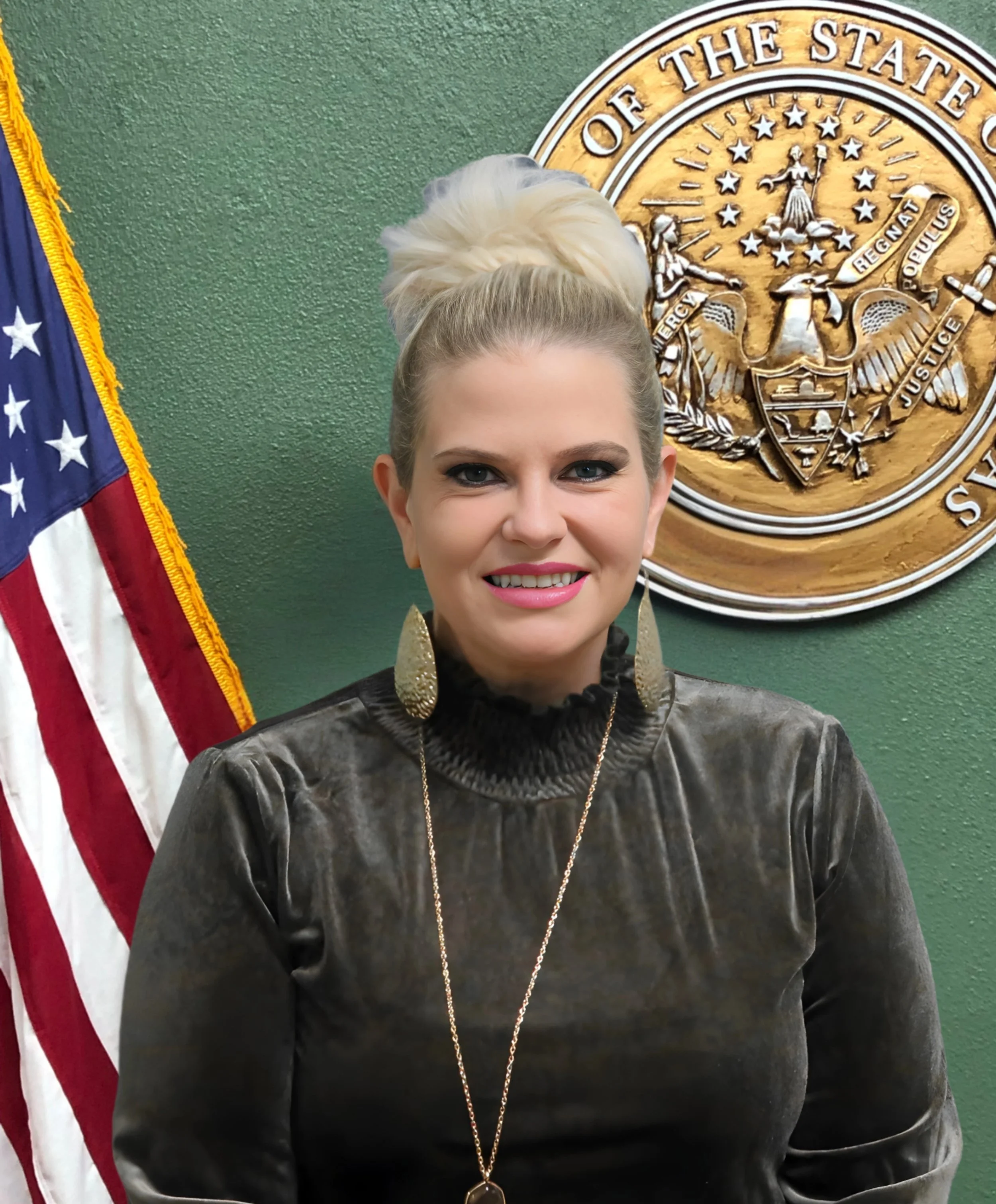 A woman with blonde hair styled in an updo, wearing gold earrings and a gold necklace, standing in front of a seal and an American flag.