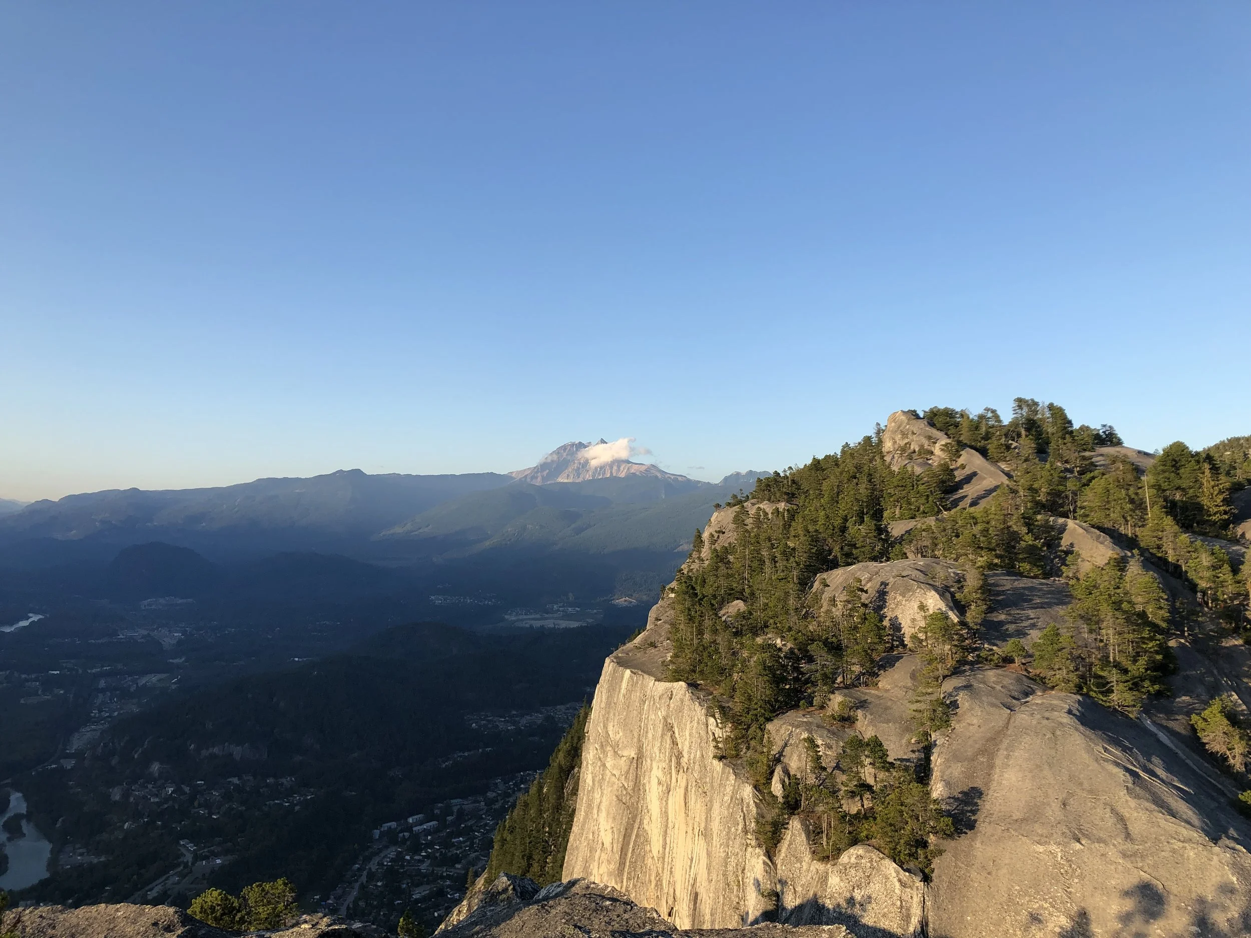 A mountainous landscape with a prominent rocky cliff on the right, covered in green pine trees. In the background, a mountain capped with snow and clouds is visible under a clear blue sky.