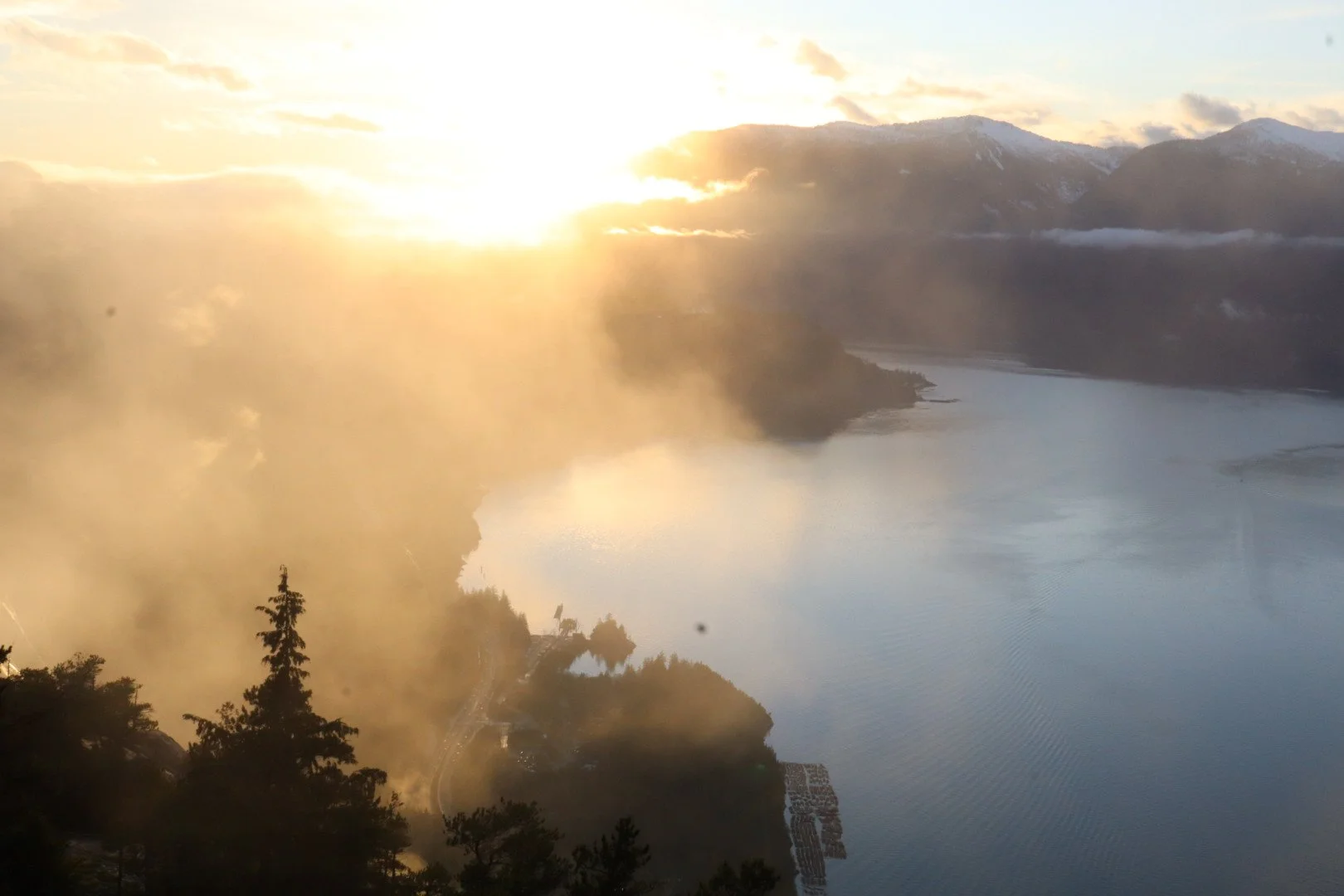 Sunrise over a body of water with mountains in the background, mist or steam on the landscape, trees in the foreground, and a few structures along the shore.