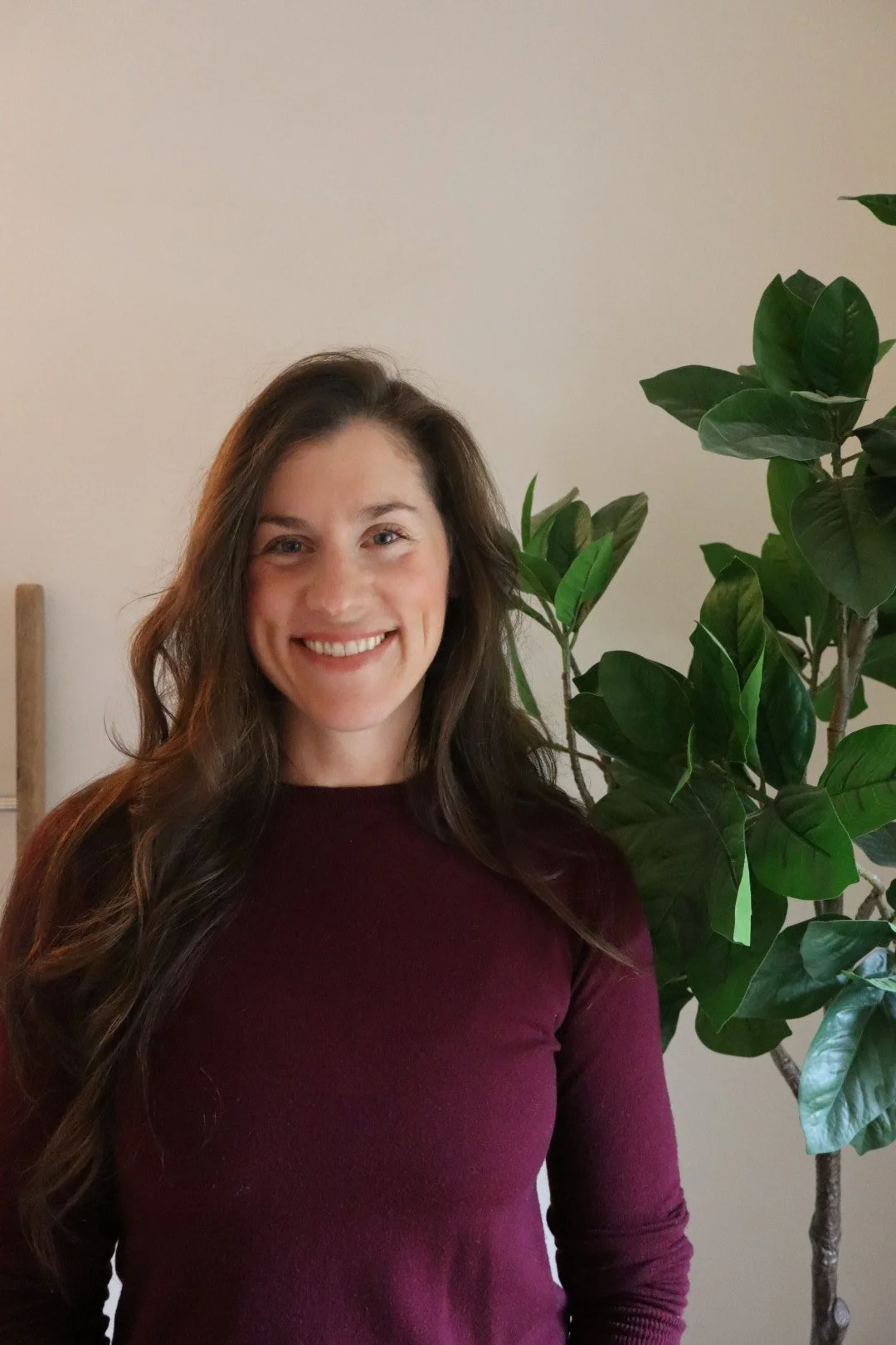 A woman with long brown hair smiling, wearing a maroon long-sleeve shirt, standing next to a large green plant with dark glossy leaves, against a plain off-white wall.