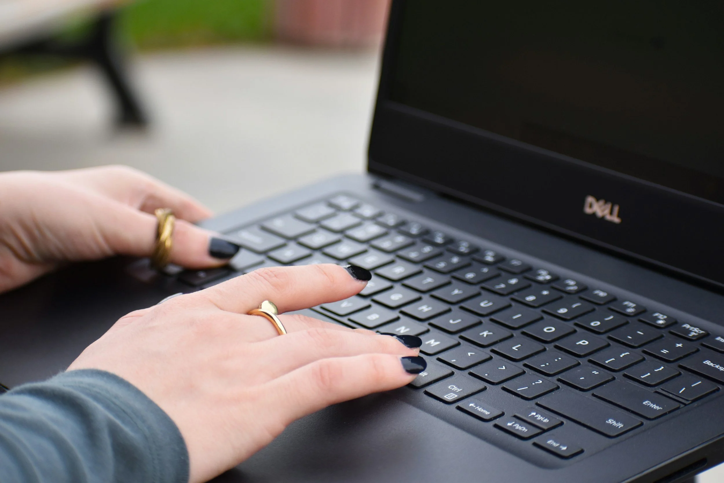 Person typing on a black Dell laptop outdoors, wearing rings on fingers and dark nail polish.