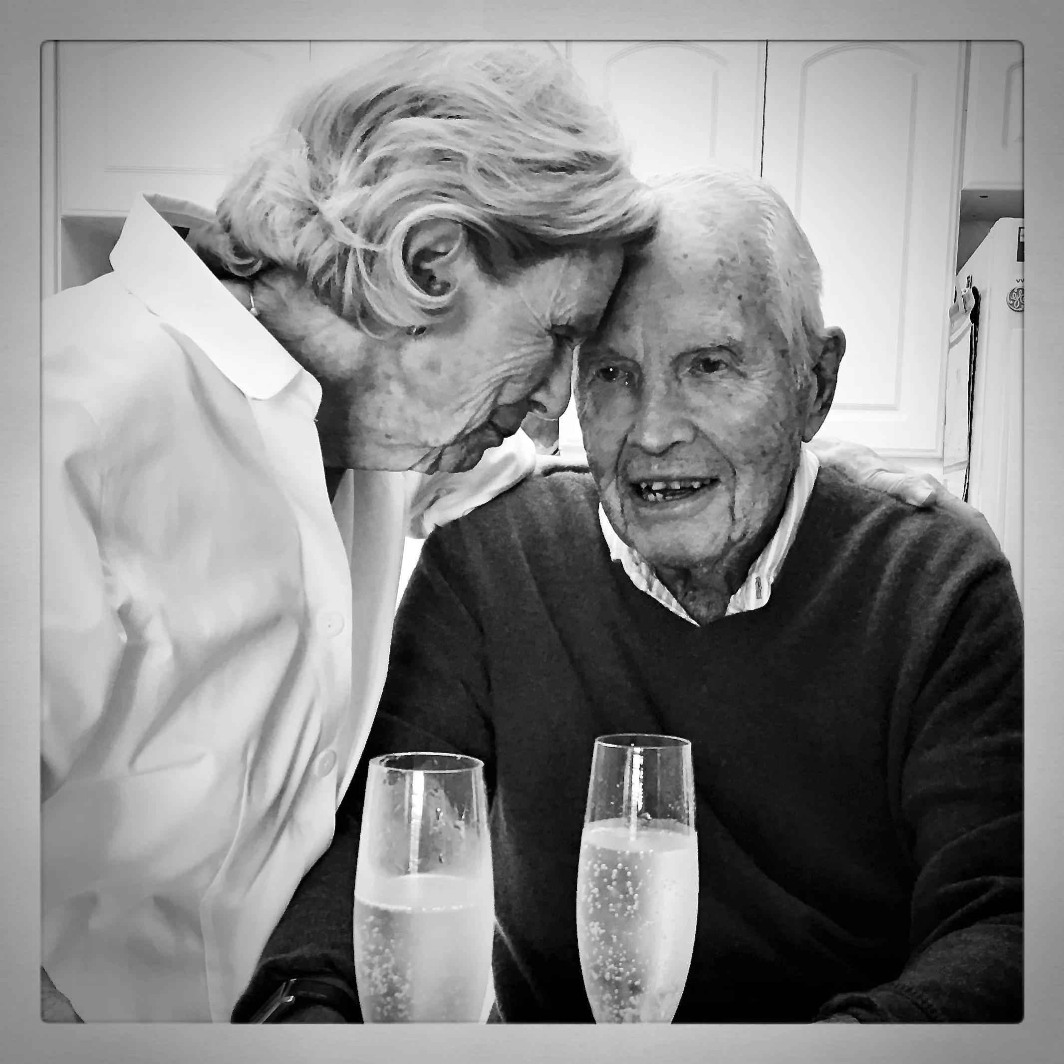 An elderly woman with curly hair is leaning her forehead against the forehead of an elderly man with thinning hair, both smiling. They are sitting at a table with two glasses of champagne in front of them, in a kitchen setting.