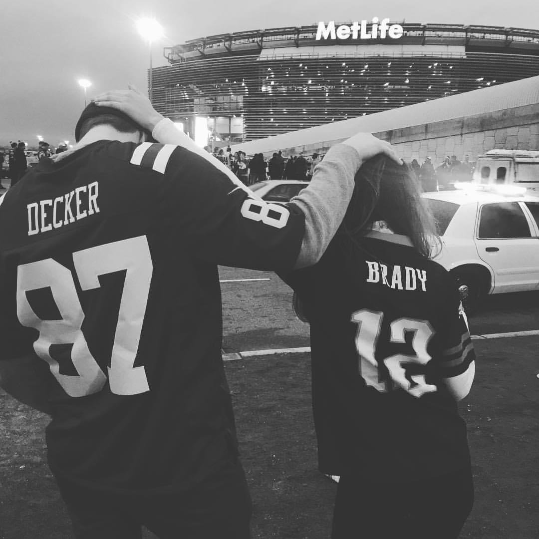 Two people wearing football jerseys, one with the name Decker and the other with the name Brady, stand outside with their arms around each other's shoulders during an evening at MetLife stadium.