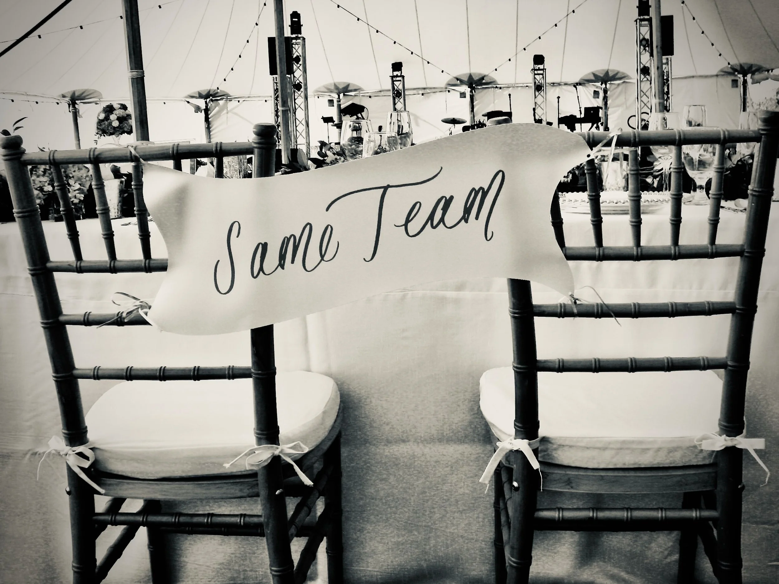 Two wooden chairs with white cushions at a wedding event attached together with a 'Same Team' banner, inside a large tent with tables decorated with flowers, and musical instruments in the background.