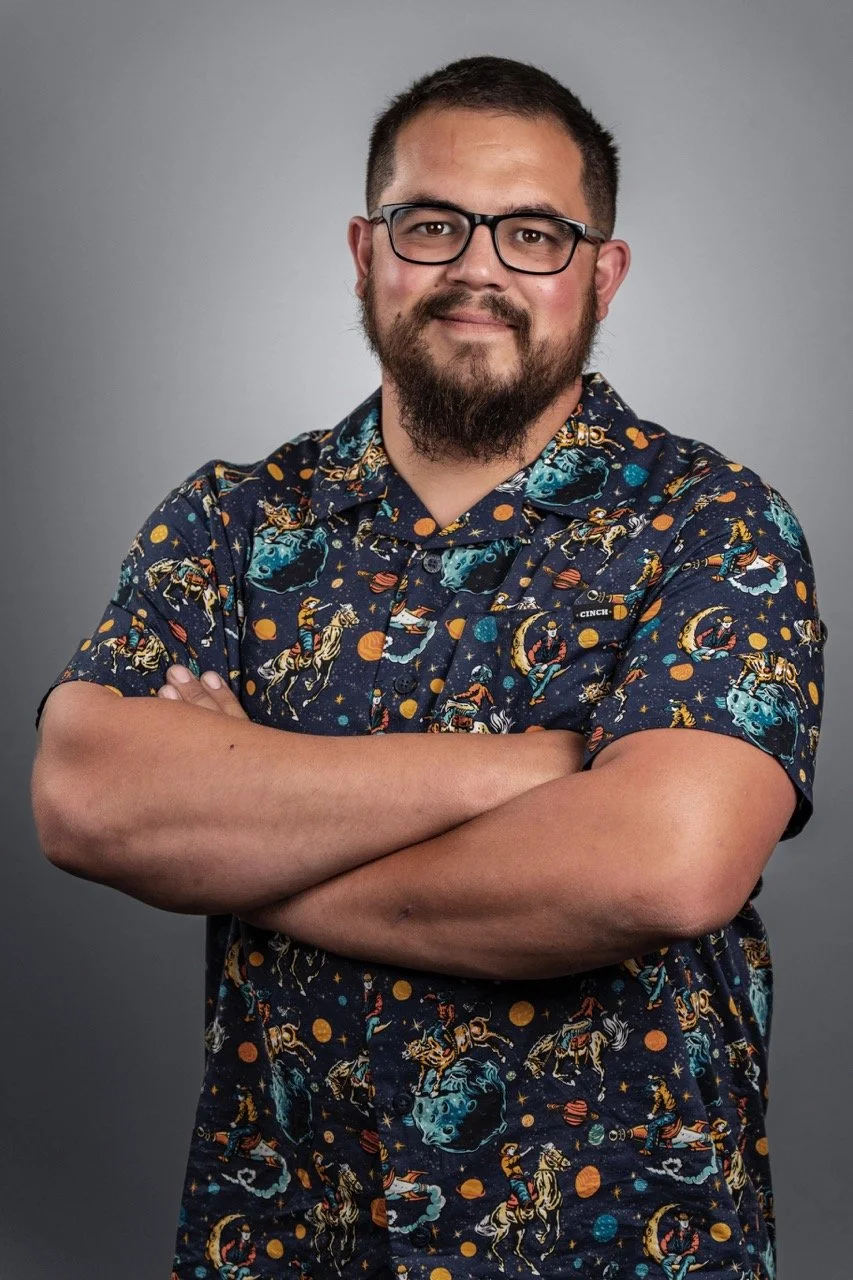 A man with glasses and a beard, wearing a colorful celestial-themed shirt, standing with arms crossed against a gray background.